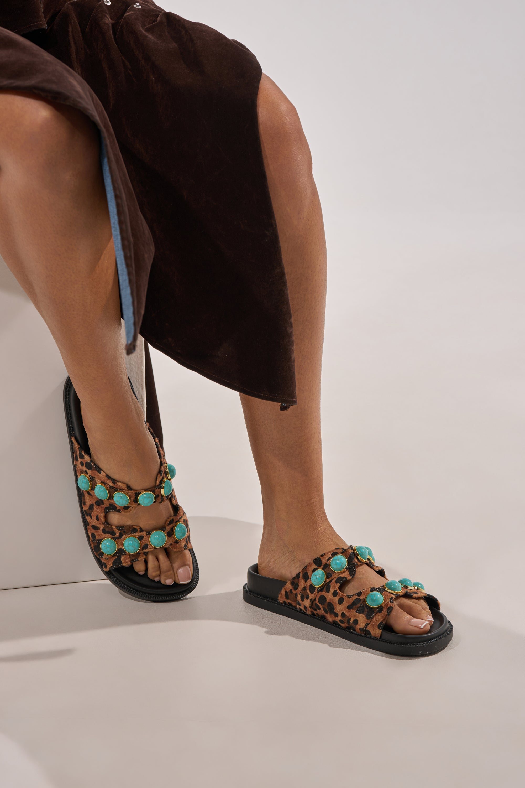 A person in a brown skirt sits cross-legged, highlighting the AZALEA WANG UZUMATI LEOPARD SLIP ON SANDAL WITH STONES—leopard print calf hair slides featuring turquoise stones on black soles. 0