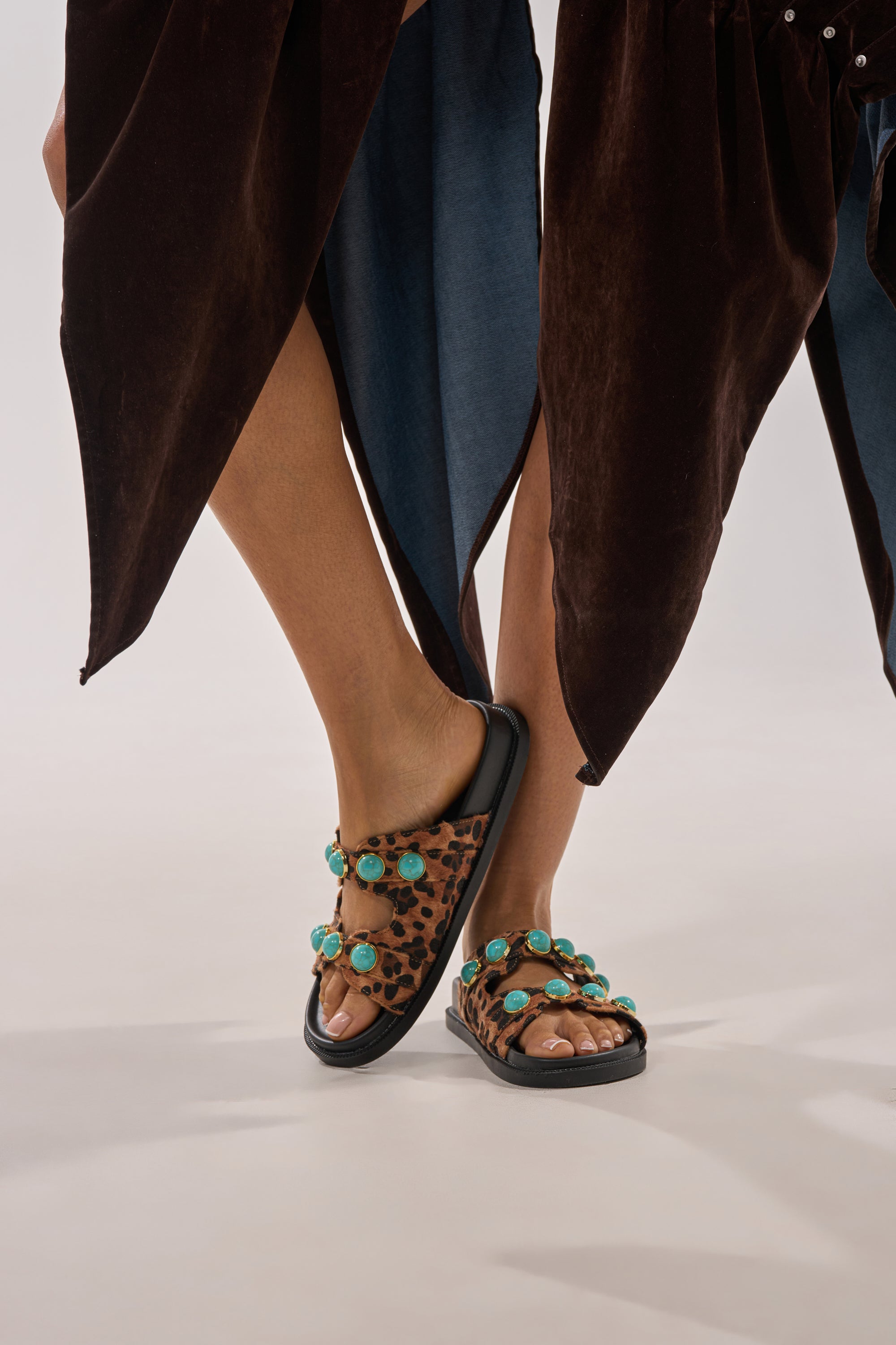 A person in a flowy brown skirt stands with crossed legs, showcasing the AZALEA WANG UZUMATI Leopard Slip On Sandal with turquoise stone embellishments against a plain light background. 1
