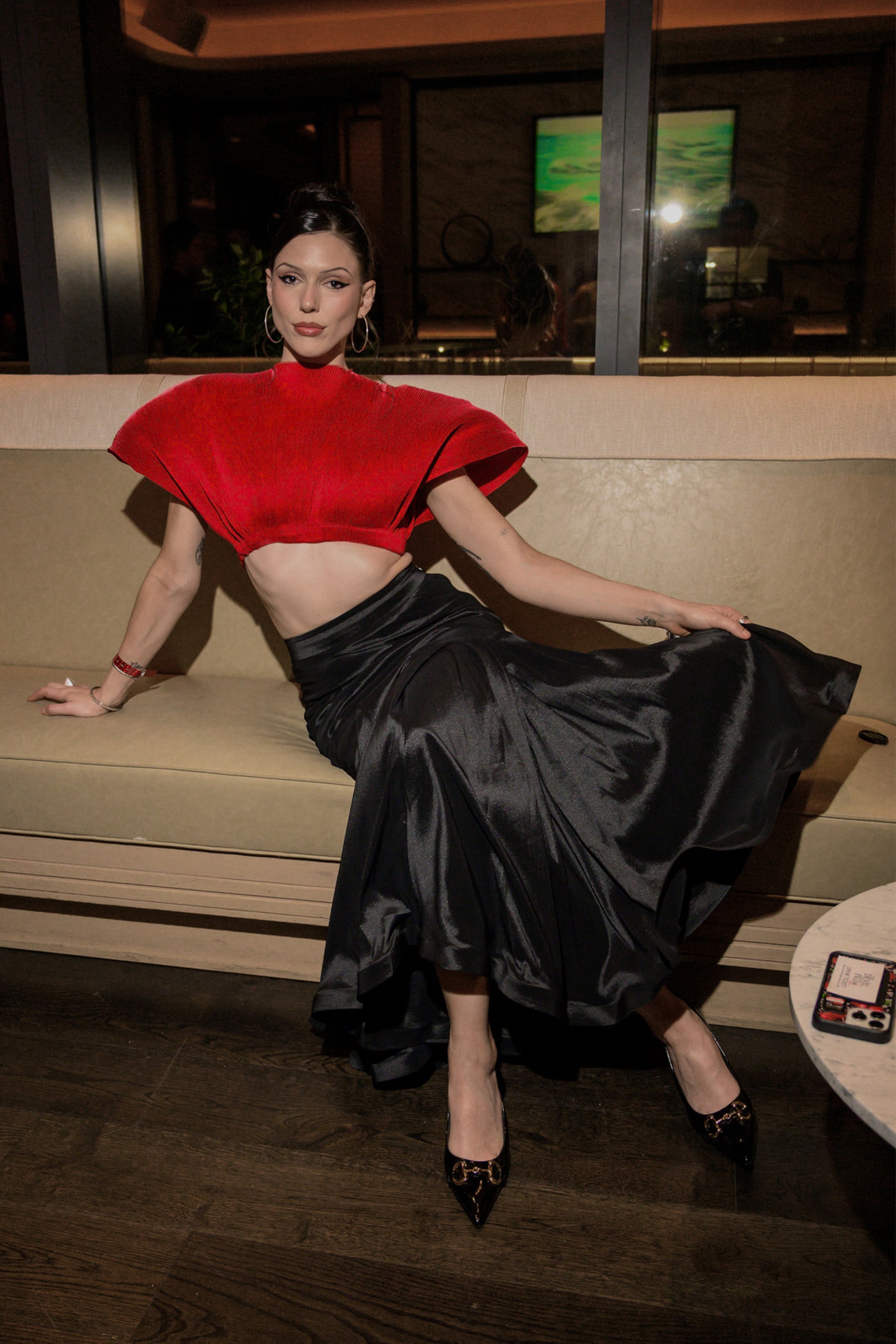 A woman poses on a beige sofa in the ALL ABOUT THE DRAMA TOP, featuring sculptural shoulders, paired with a flowing black satin skirt and black heels. The upscale modern lounge enhances her statement fashion look. 1