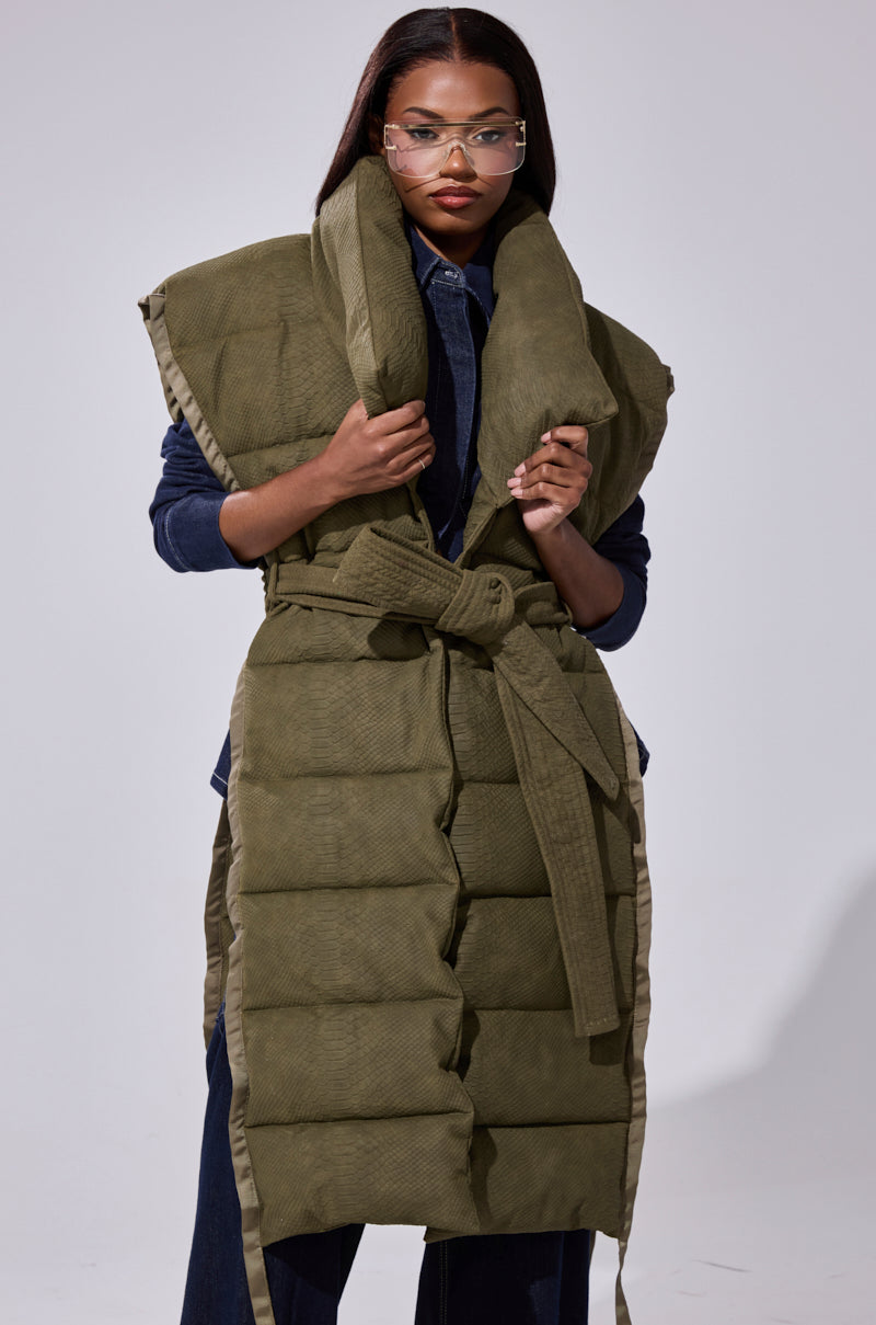 A woman wearing glasses and the PILOT JONES LONGLINE PUFFER VEST IN CROC SUEDE, featuring an oversized collar and tied waist, over a dark denim outfit. She stands against a plain light background.