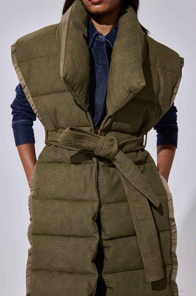 A person in a dark denim shirt wears the PILOT JONES LONGLINE PUFFER VEST IN CROC SUEDE, featuring an olive-green hue, oversized collar, and matching fabric belt. The face is mostly out of frame.