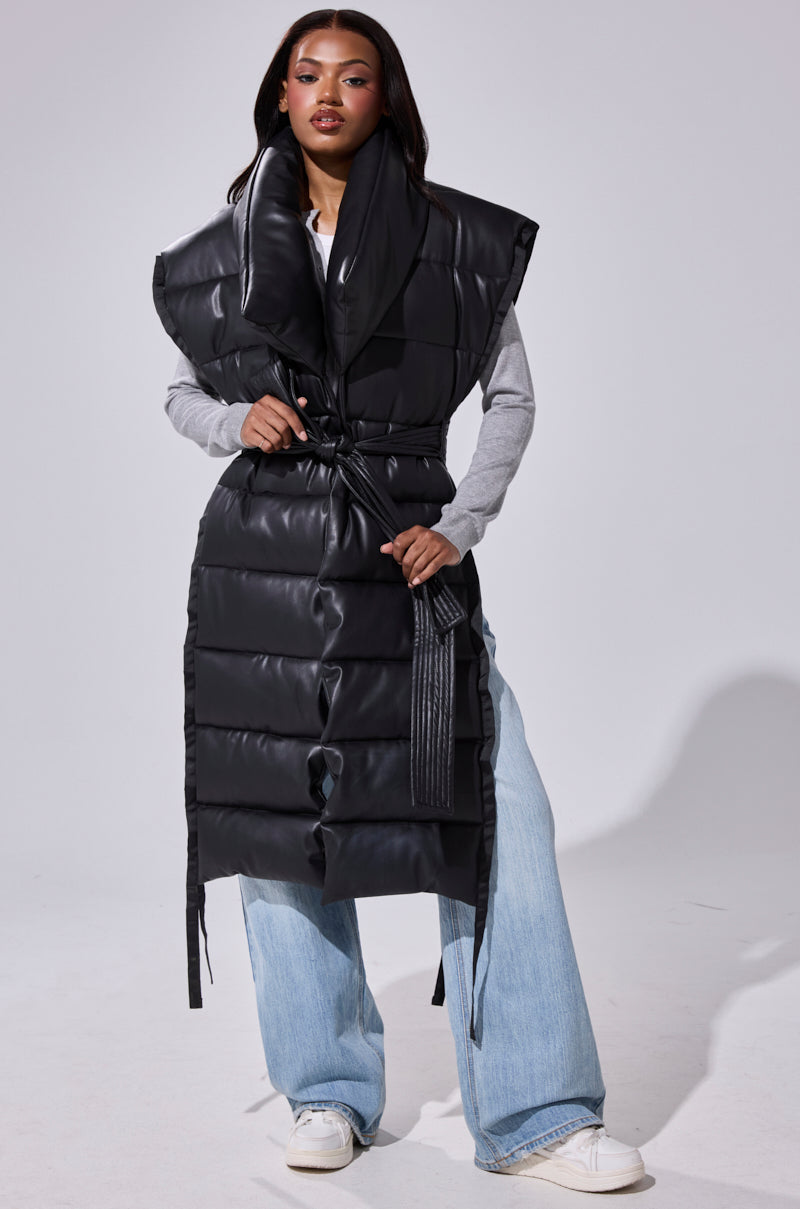 A woman wears the PILOT JONES Longline Puffer Vest in faux leather with an oversized collar and belted waist over a gray top, wide-leg light blue jeans, and white sneakers.