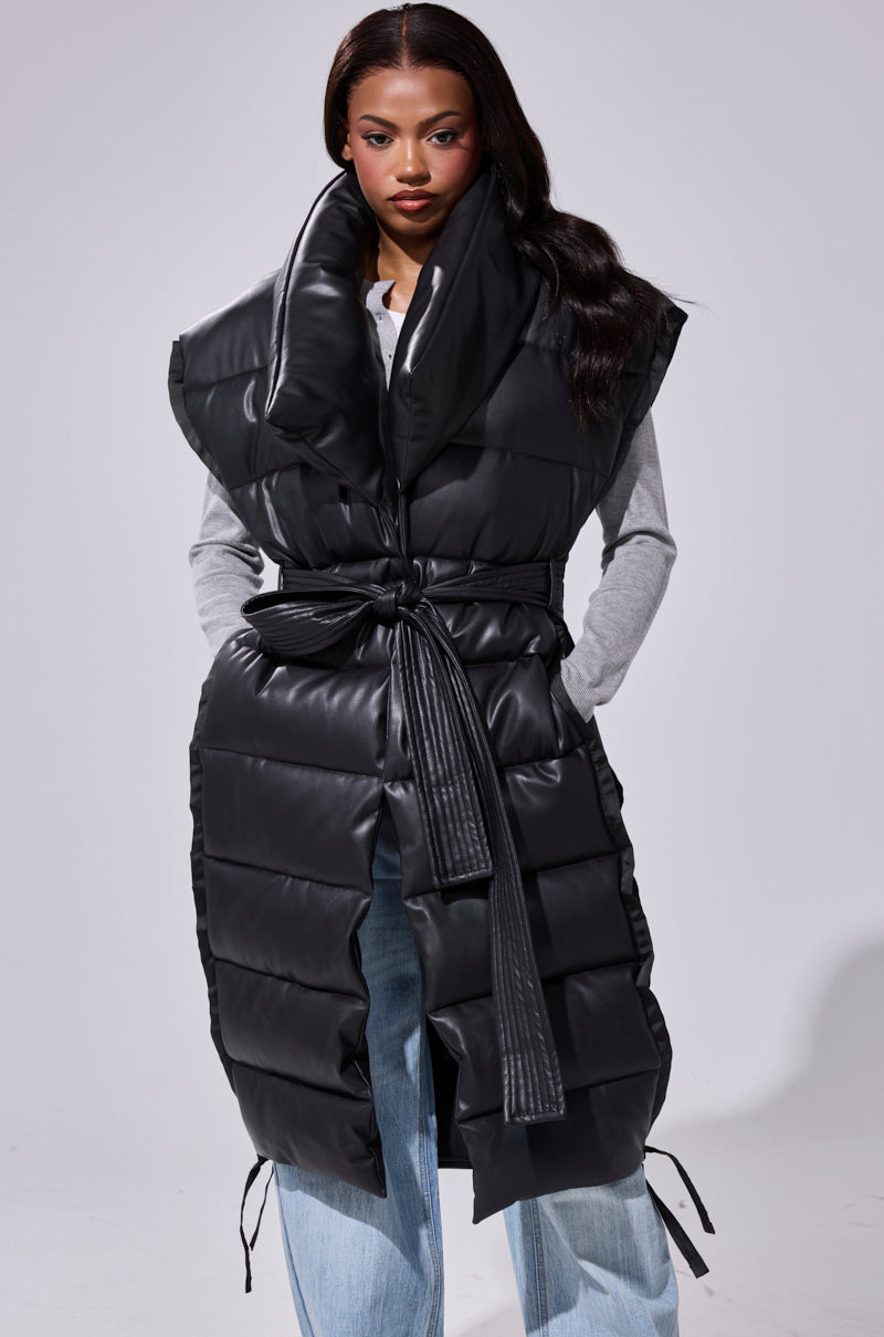 A woman stands facing forward against a plain light background, wearing the PILOT JONES Longline Puffer Vest in faux leather with an oversized collar over a gray long-sleeve top and light blue jeans.