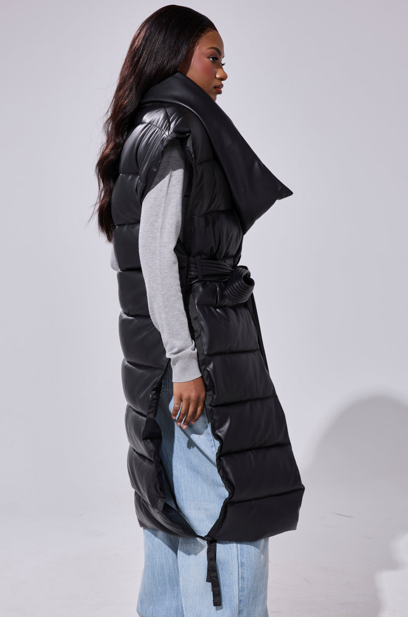 A woman stands sideways against a plain light background, wearing the PILOT JONES LONGLINE PUFFER VEST IN FAUX LEATHER with an oversized collar over a gray long-sleeve shirt and light blue wide-leg jeans.