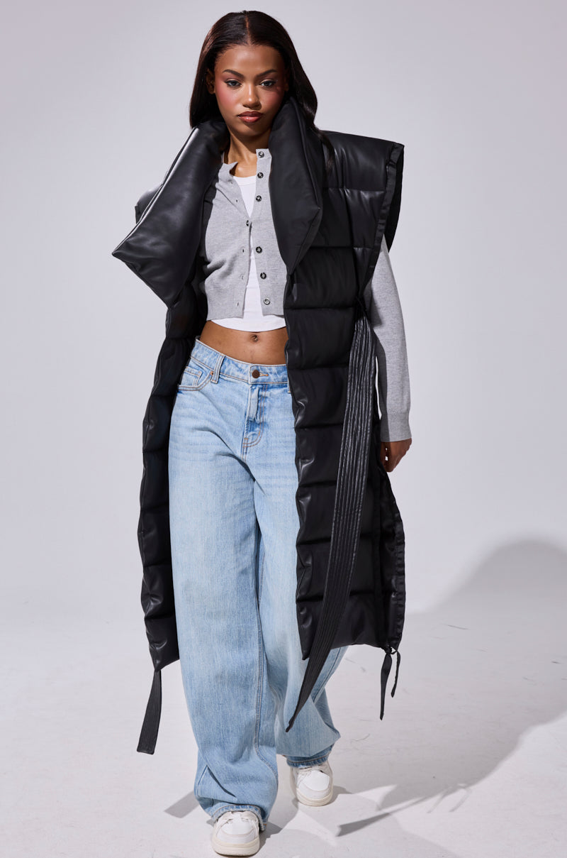 A woman models the PILOT JONES Longline Puffer Vest in Faux Leather over a cropped gray cardigan, loose light blue jeans, and white sneakers while walking confidently against a plain white background.