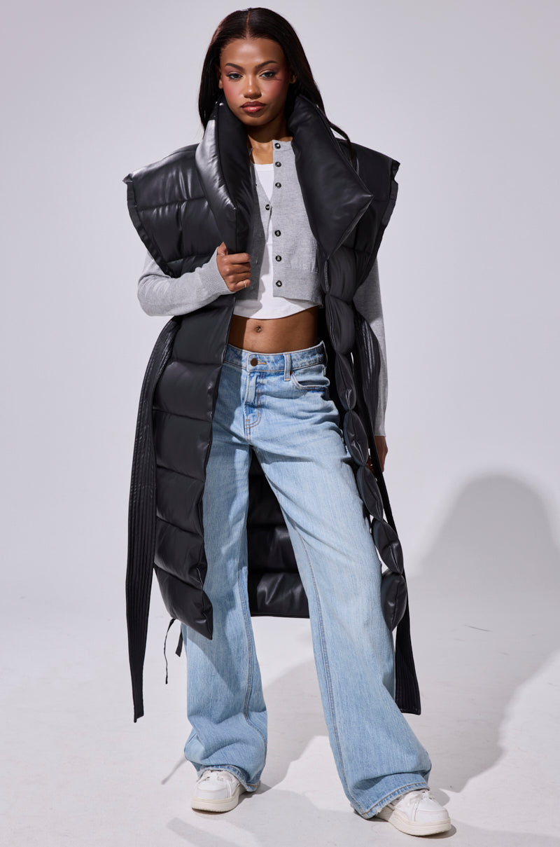 A woman with straight dark hair wears the PILOT JONES Longline Puffer Vest in Faux Leather, featuring an oversized collar, over a gray cropped henley and light blue wide-leg jeans with white sneakers. She stands facing forward with a confident expression.