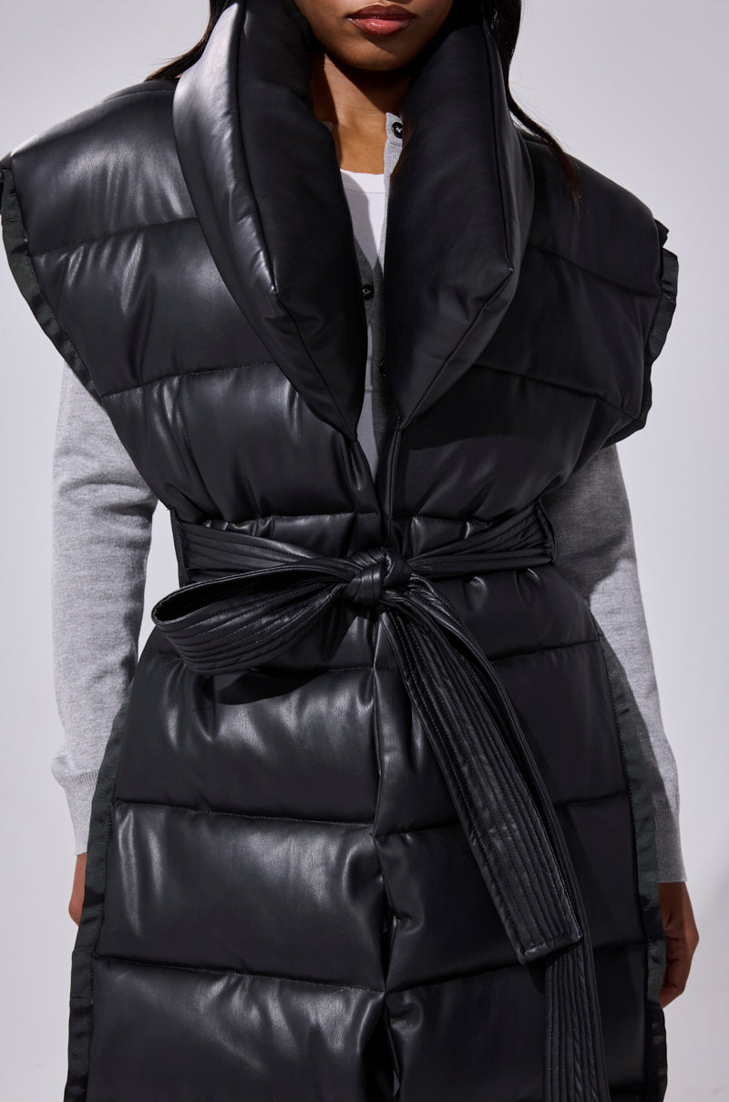 A woman wears the PILOT JONES LONGLINE PUFFER VEST IN FAUX LEATHER, a black sleeveless vest with an oversized collar and tied belt, layered over a light gray long-sleeve top. The image is cropped at her nose and knees.