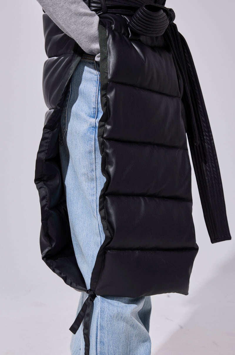 A person wears light blue jeans, a grey top, and a black quilted wrap skirt with an open side and tied belt, styled with the PILOT JONES LONGLINE PUFFER VEST IN FAUX LEATHER. Photo is cropped to show only the torso and legs.