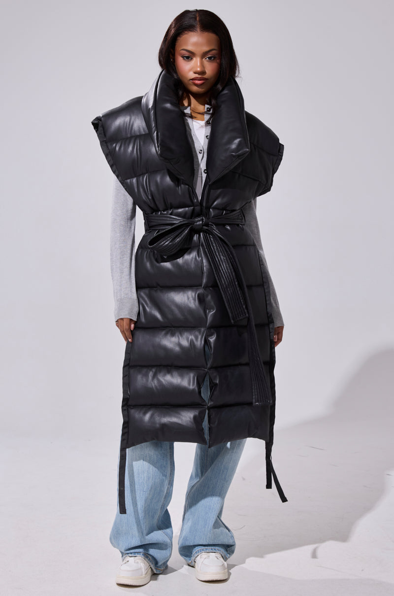 A woman faces forward in the PILOT JONES LONGLINE PUFFER VEST IN FAUX LEATHER, featuring an oversized collar and belted waist, layered over a light gray top, light blue jeans, and white sneakers against a plain light background.