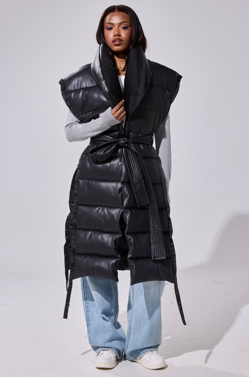 A person wears the PILOT JONES LONGLINE PUFFER VEST IN FAUX LEATHER with an oversized collar and tie belt over a gray long-sleeve top, light blue jeans, and white sneakers, standing against a plain light background.