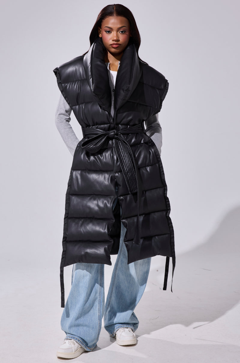 A woman wears the PILOT JONES Longline Puffer Vest in Faux Leather—black, sleeveless, belted with an oversized collar—over a gray long-sleeve shirt and light blue wide-leg jeans, white sneakers completing the look against a plain light background.