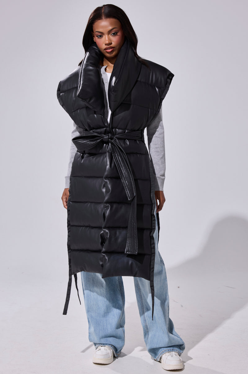 A woman stands against a white background wearing the PILOT JONES Longline Puffer Vest in Faux Leather with an oversized collar and tie belt, layered over a gray sweater, light blue jeans, and white sneakers.