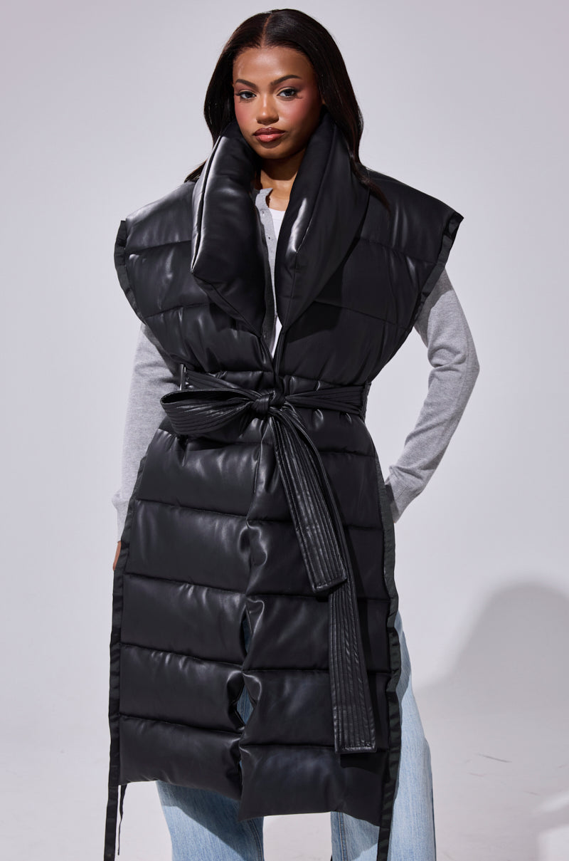 A woman models the PILOT JONES LONGLINE PUFFER VEST IN FAUX LEATHER with an oversized collar and tied belt, layered over a light gray long-sleeve shirt and blue jeans against a plain background.