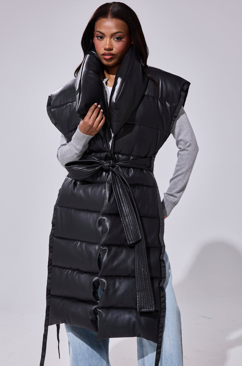 A woman wears the PILOT JONES Longline Puffer Vest in Faux Leather with an oversized collar and tied belt over a light gray long-sleeve shirt and light blue jeans, standing against a plain background.