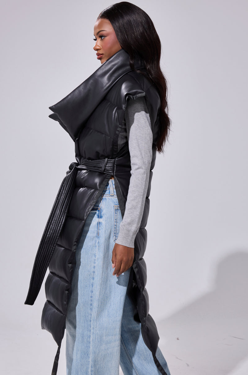 A woman stands sideways in the PILOT JONES LONGLINE PUFFER VEST IN FAUX LEATHER, featuring a black belted design and oversized collar, layered over a gray long-sleeve top and light blue wide-leg jeans against a plain light background.