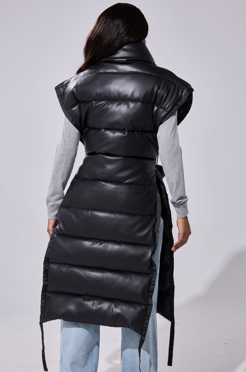 A person faces away wearing the PILOT JONES Longline Puffer Vest in Faux Leather with cap sleeves over a gray long-sleeve shirt and light blue jeans. The quilted vest has an oversized collar and falls below the knees.