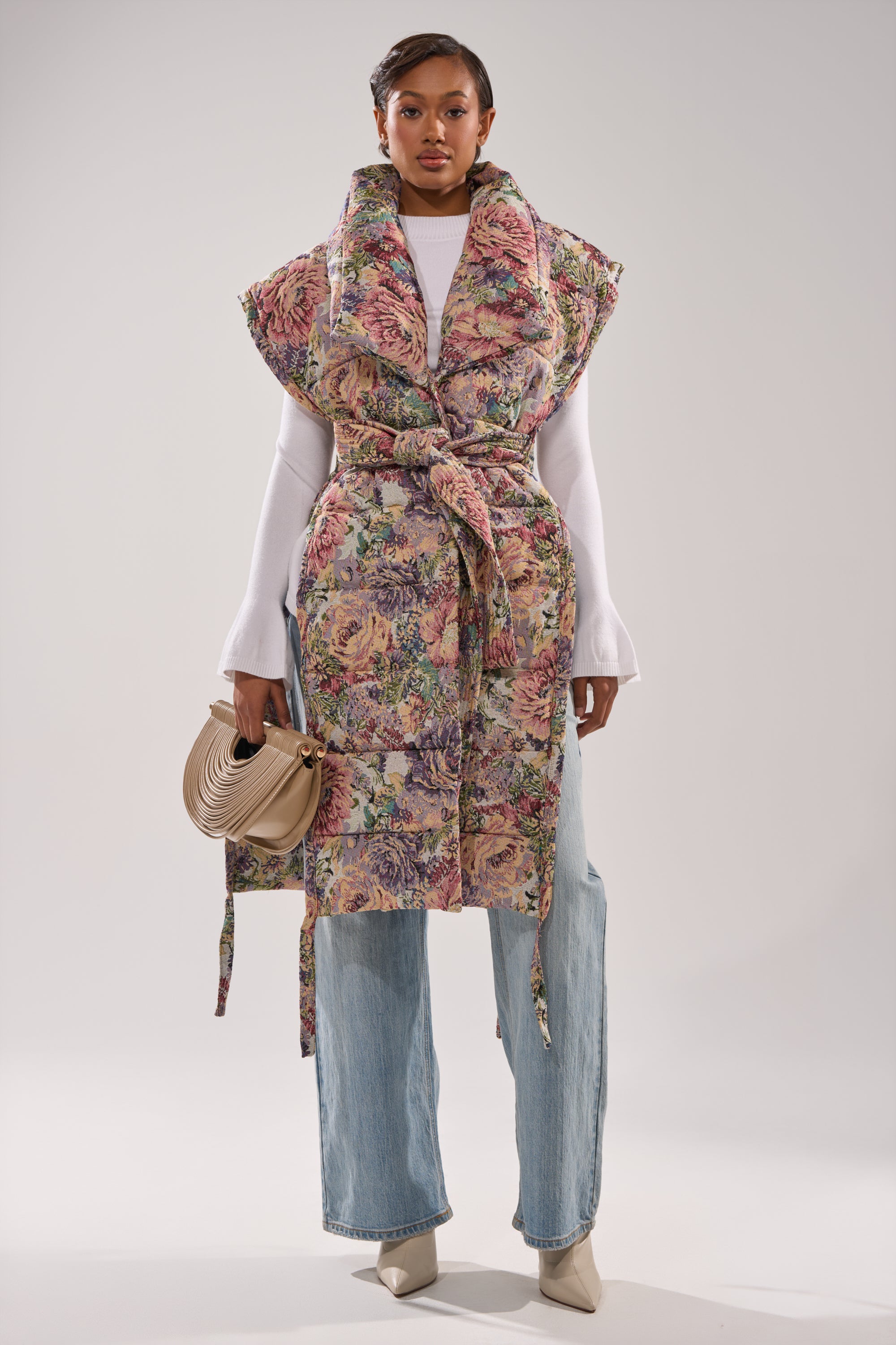 A woman wears the PILOT JONES LONGLINE PUFFER VEST IN TAPESTRY, styled with a white long-sleeve top, light blue wide-leg jeans, beige heeled boots, and a beige purse against a plain light background.