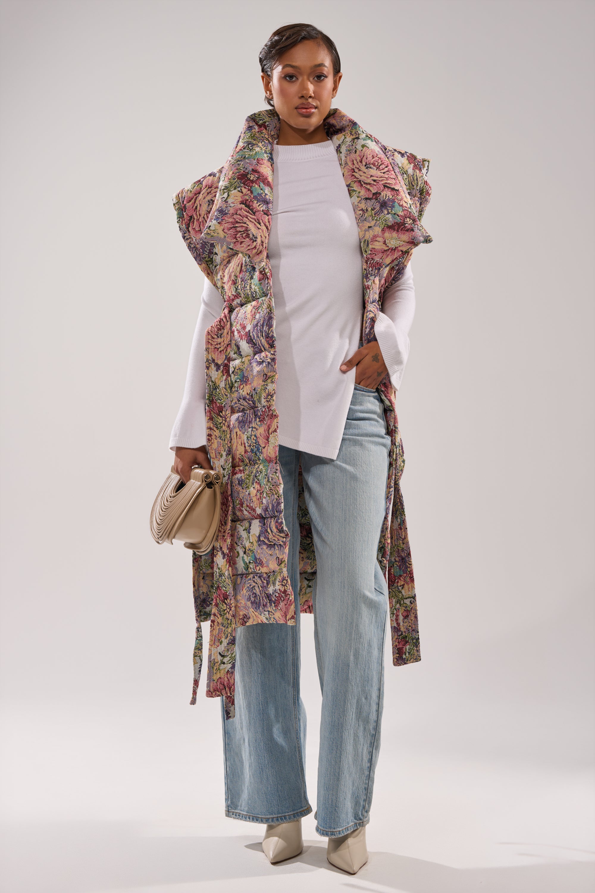 A woman styles the PILOT JONES LONGLINE PUFFER VEST IN TAPESTRY over a white long-sleeve top, light blue wide-leg jeans, and cream heels, holding a beige handbag against a plain background.