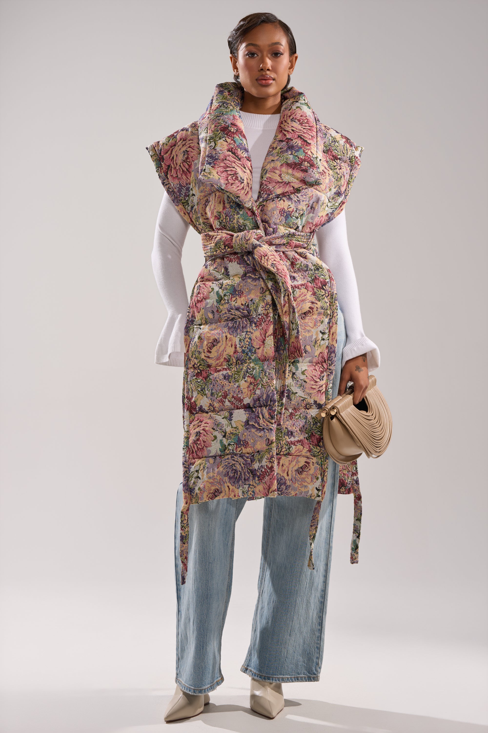 A woman wears the PILOT JONES LONGLINE PUFFER VEST IN TAPESTRY over a white turtleneck, paired with light blue wide-leg jeans, beige pointed-toe boots, and carries a small beige purse.