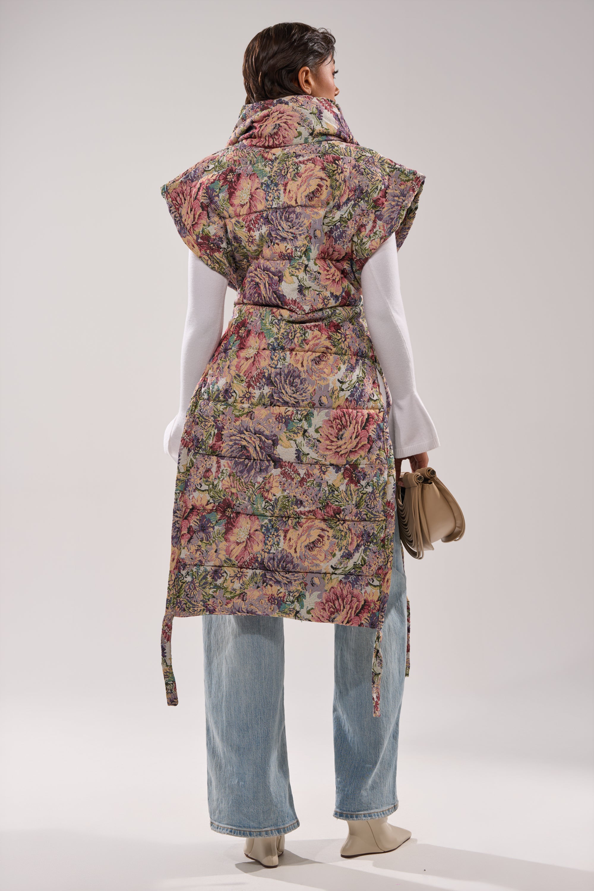 A person stands facing away, wearing the PILOT JONES LONGLINE PUFFER VEST IN TAPESTRY over a white top and light blue jeans, paired with a small beige handbag and cream-colored heeled shoes.