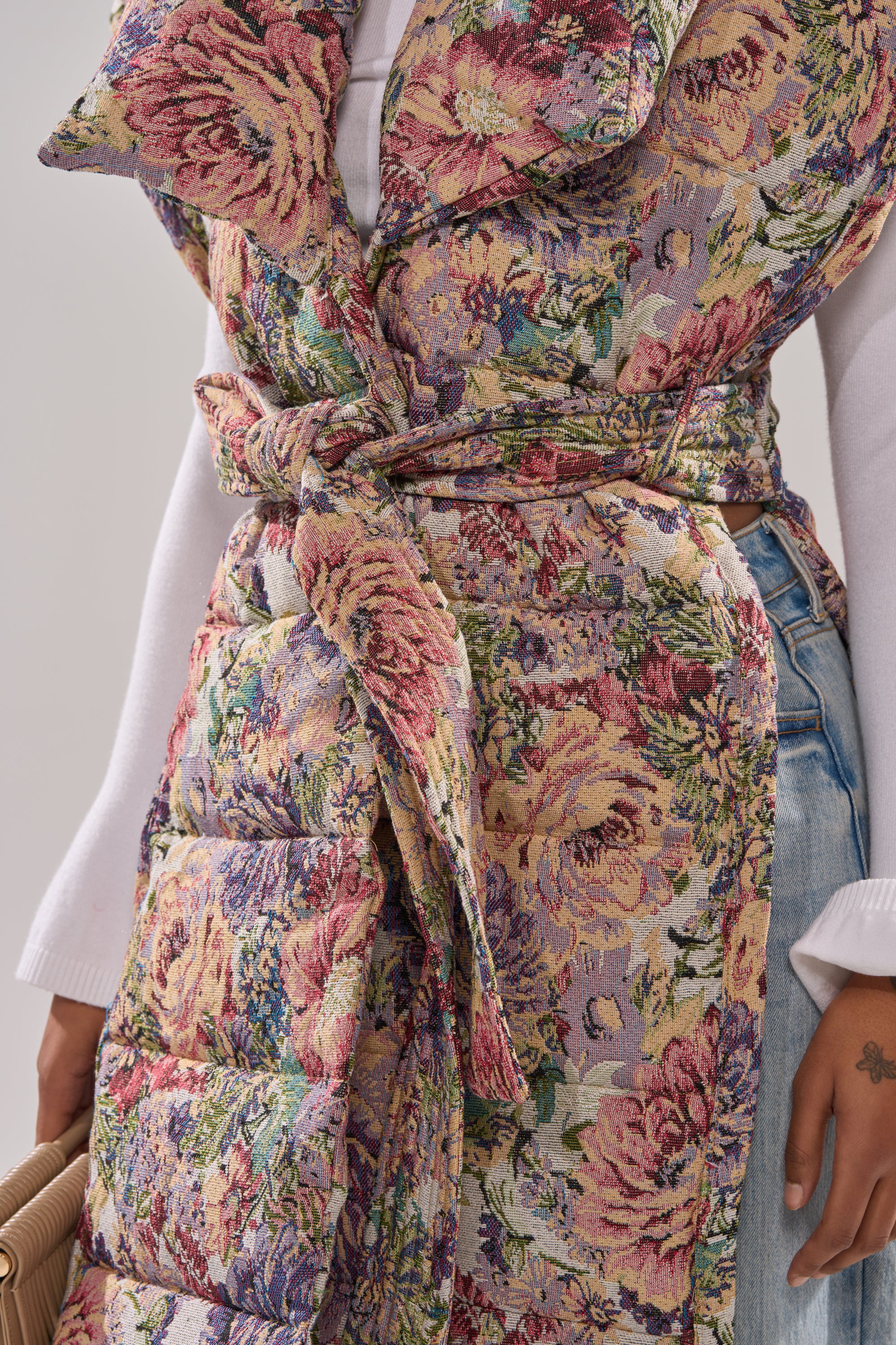 A person wearing the PILOT JONES LONGLINE PUFFER VEST IN TAPESTRY over a white long-sleeve top and light blue jeans, holding a tan handbag.