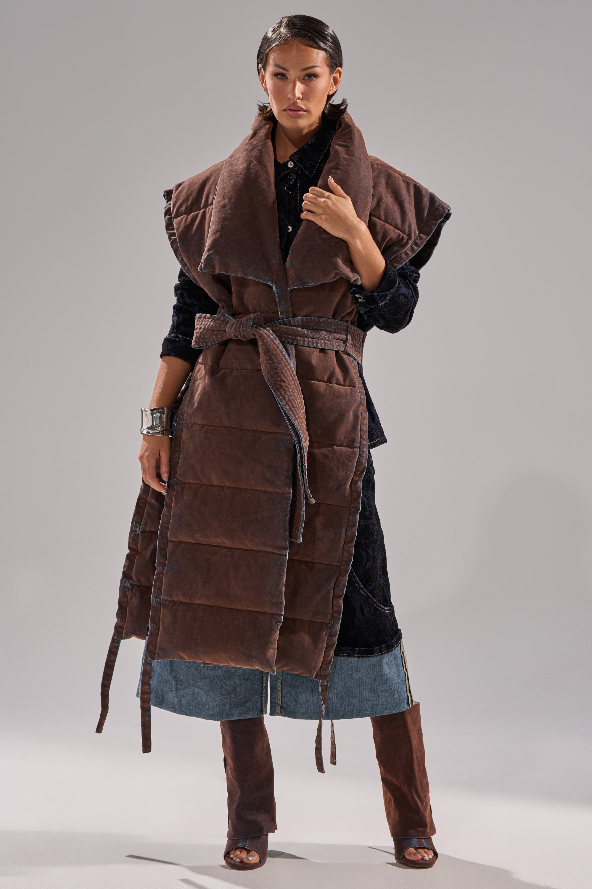 A woman stands confidently in the PILOT JONES LONGLINE PUFFER VEST IN VELVET DENIM, belted at the waist and layered over a dark textured dress, denim pants with brown suede chaps, statement silver cuffs, and open-toed boots.