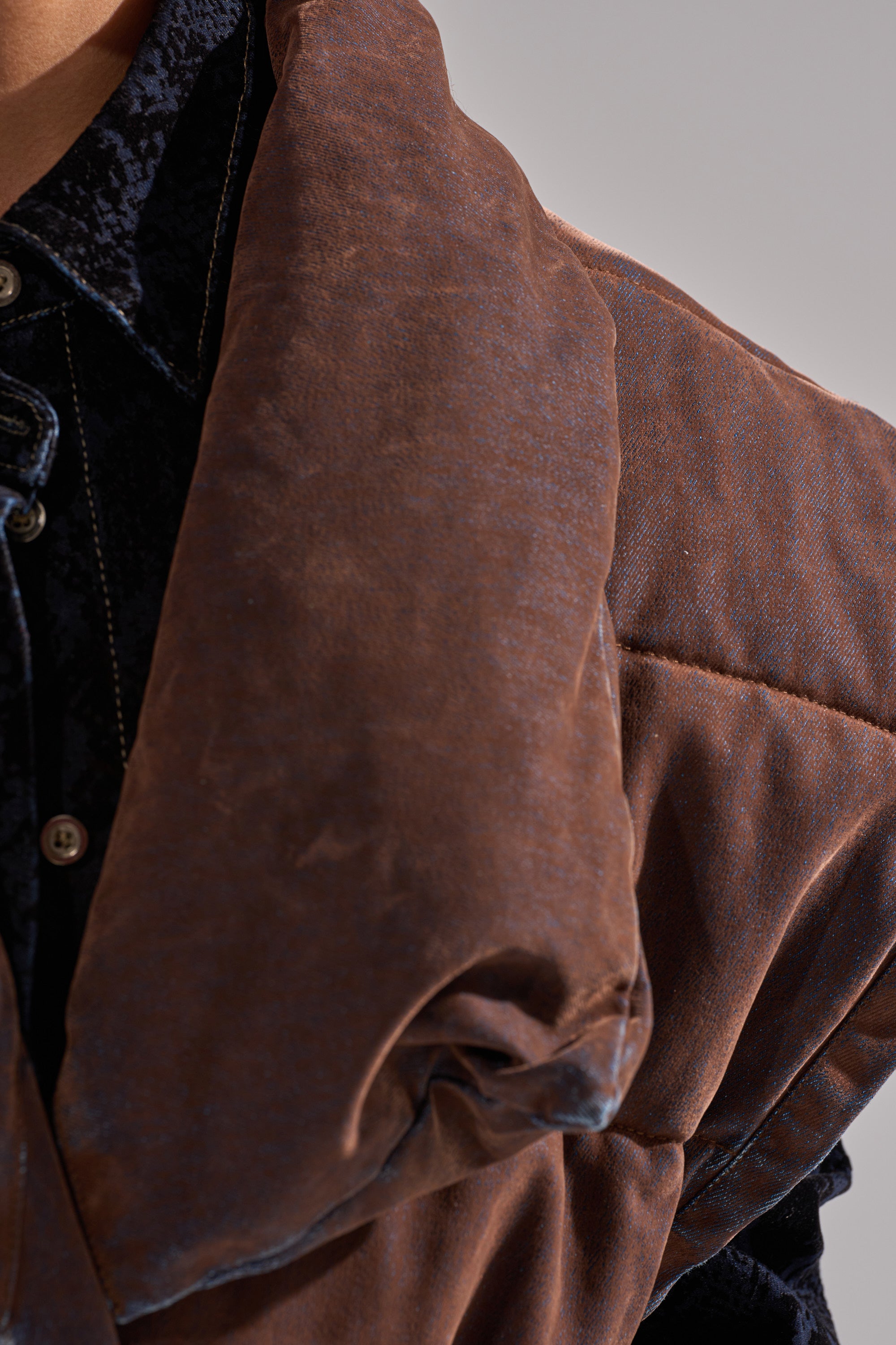 Close-up of a person wearing a dark textured shirt layered with the PILOT JONES LONGLINE PUFFER VEST IN VELVET DENIM. The focus is on the rich fabrics and layering, showing only part of the neck and shoulder.