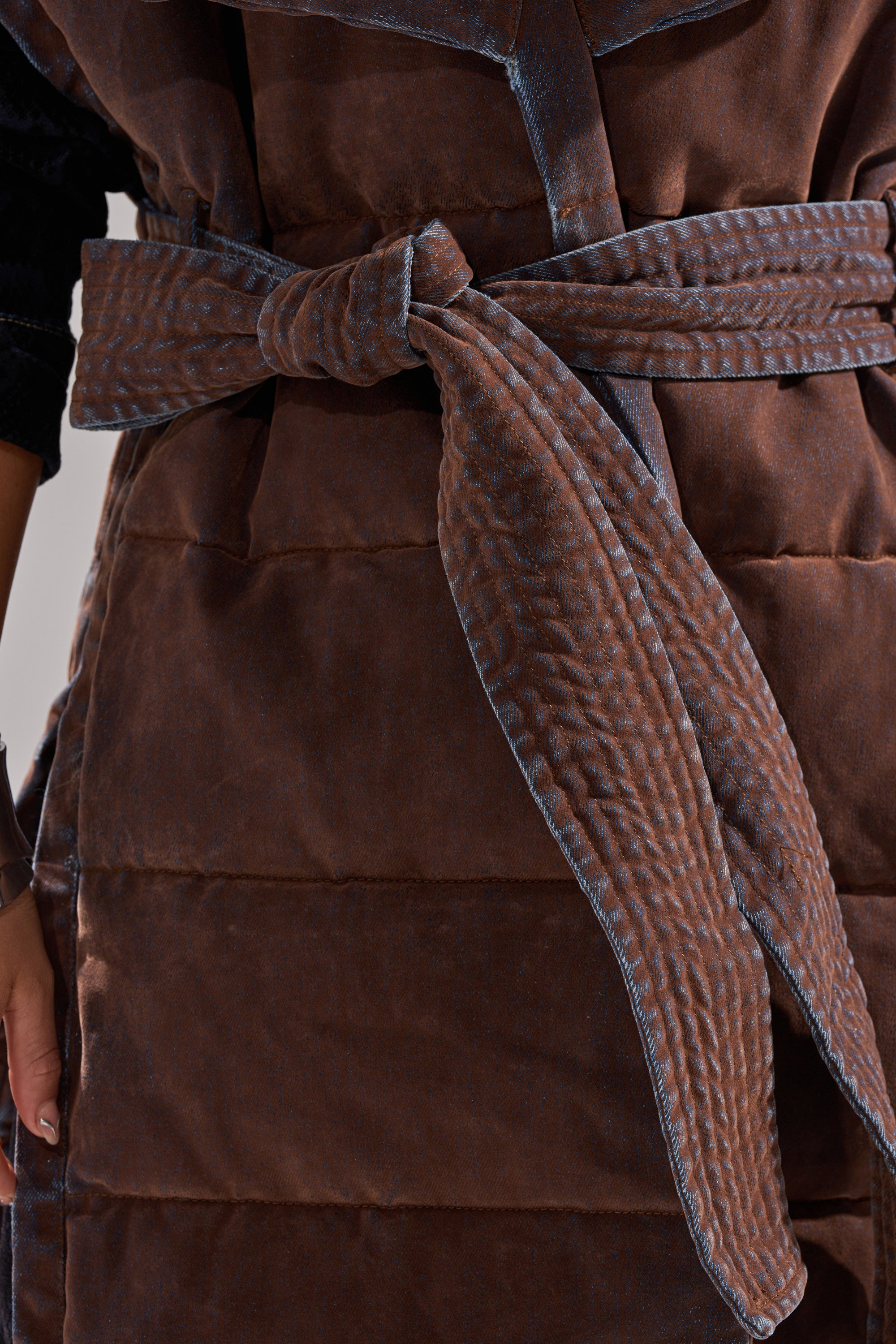 Close-up of a person wearing the PILOT JONES LONGLINE PUFFER VEST IN VELVET DENIM, featuring a quilted texture, with a blue-stitched belt tied at the waist. The person’s left hand rests partially visible at their side.