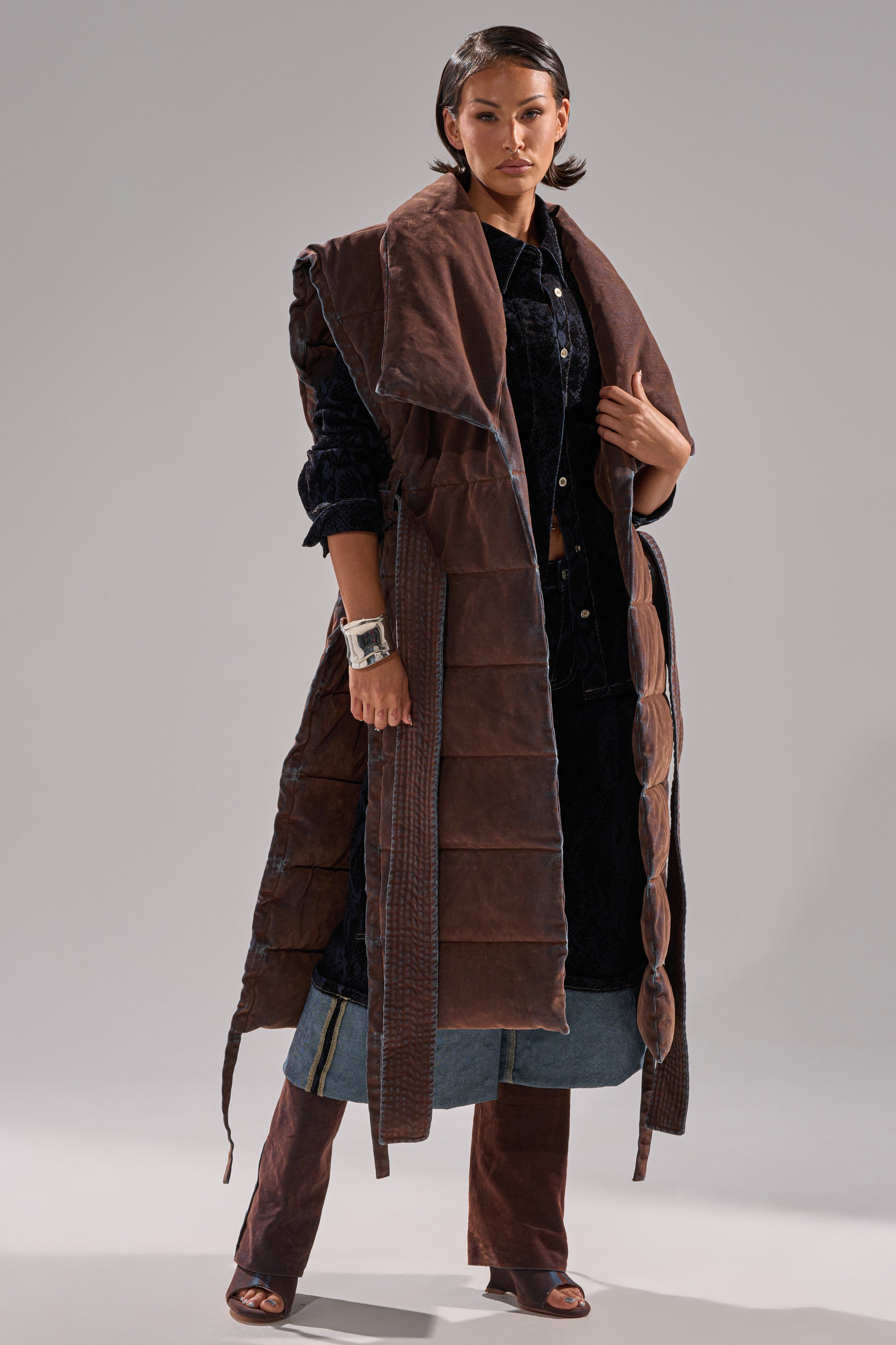 A woman stands confidently against a gray background wearing the PILOT JONES LONGLINE PUFFER VEST IN VELVET DENIM over a black top with wide sleeves, a layered denim skirt, and brown flared pants.