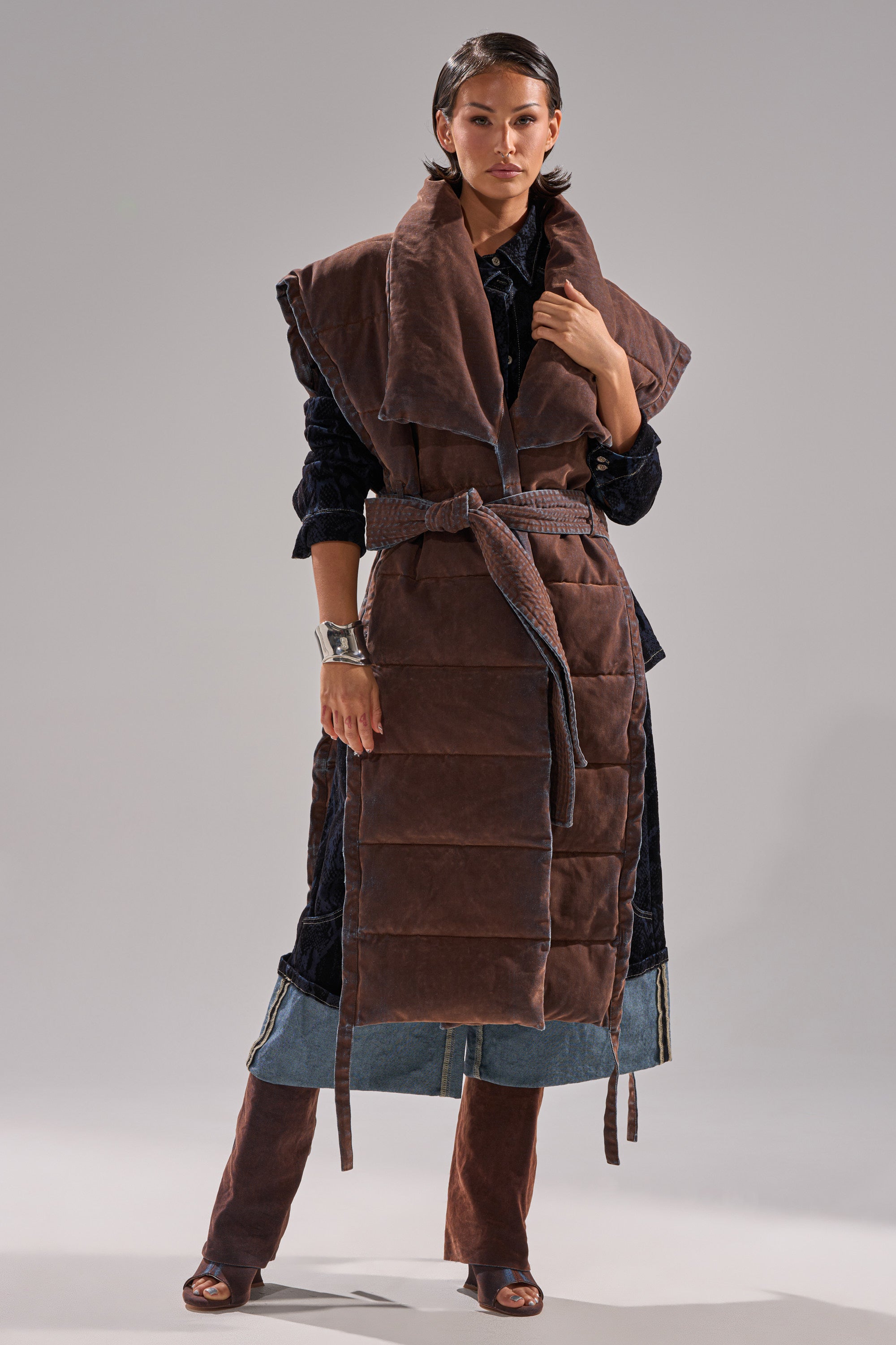 A woman, with short, slicked-back hair, stands against a plain background wearing the PILOT JONES LONGLINE PUFFER VEST IN VELVET DENIM, styled over a dark outfit with denim details and brown open-toe boots.