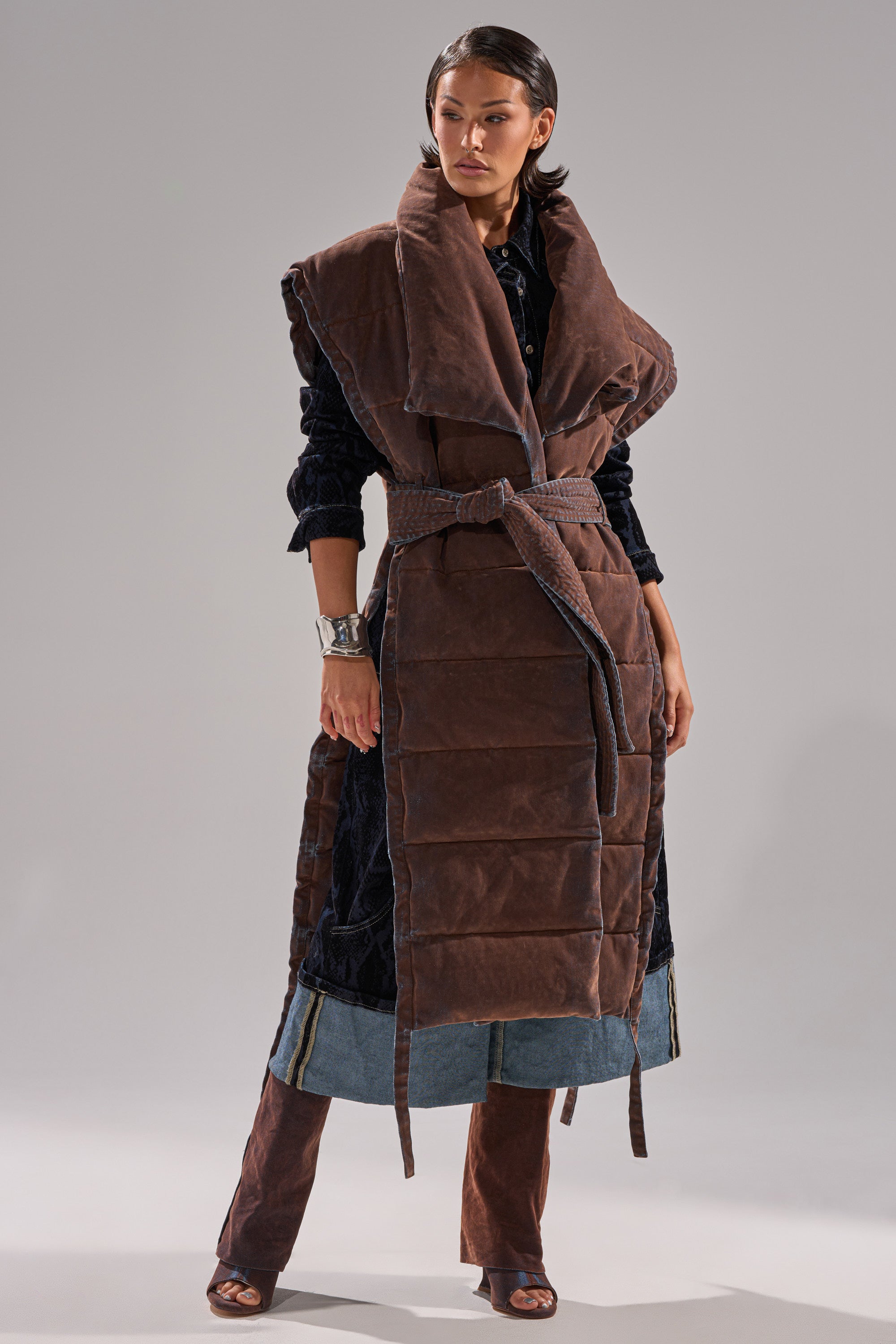 A woman wears the PILOT JONES LONGLINE PUFFER VEST IN VELVET DENIM, a long, brown padded sleeveless coat with a large collar and belted waist, layered over a dark textured outfit and denim-trimmed pants. She accessorizes with sleek hair and a silver cuff.