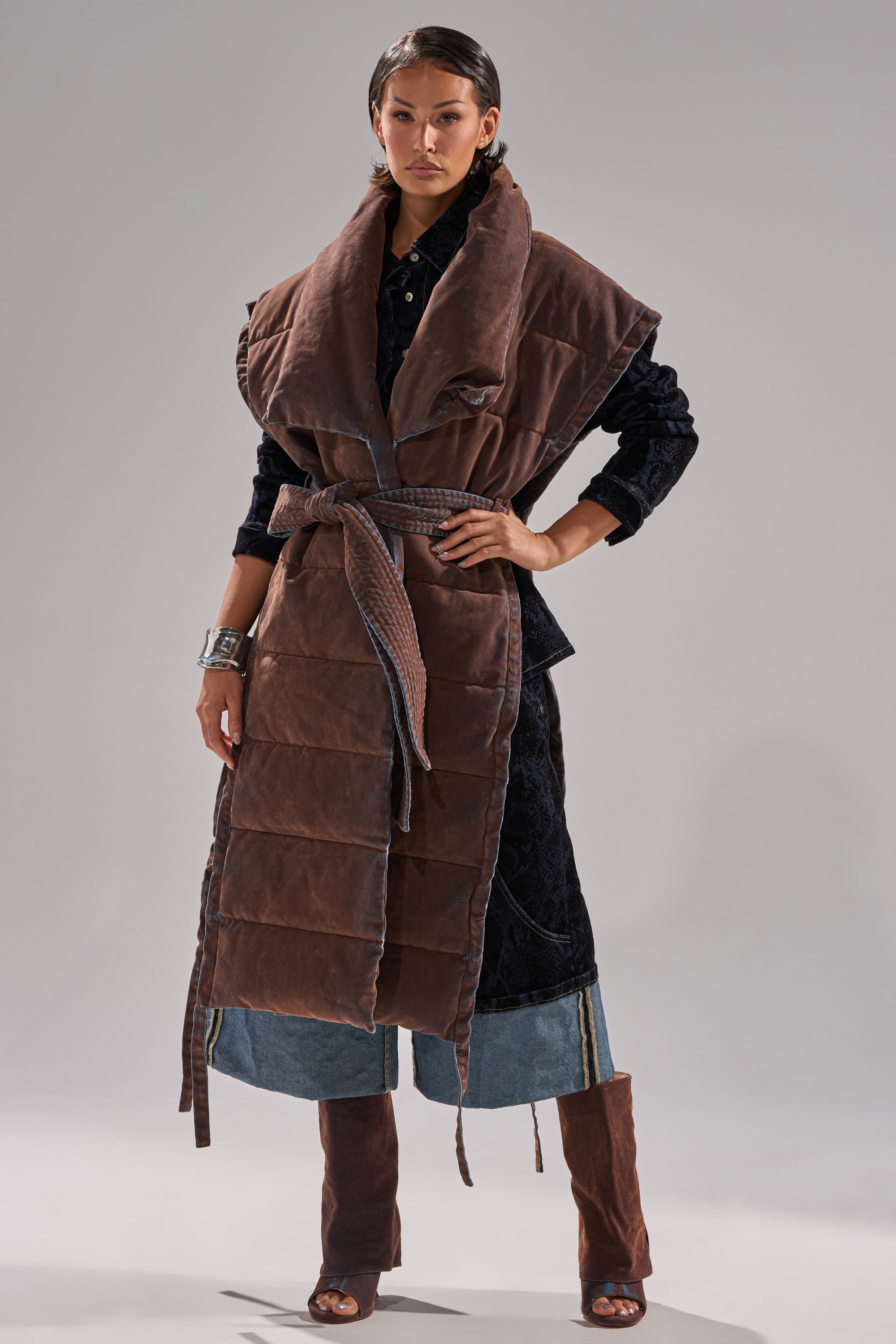 A woman stands confidently in the PILOT JONES LONGLINE PUFFER VEST IN VELVET DENIM, styled with a wide collar, belted waist, layered over a dark coat, with flared jeans and brown knee-high open-toe boots.