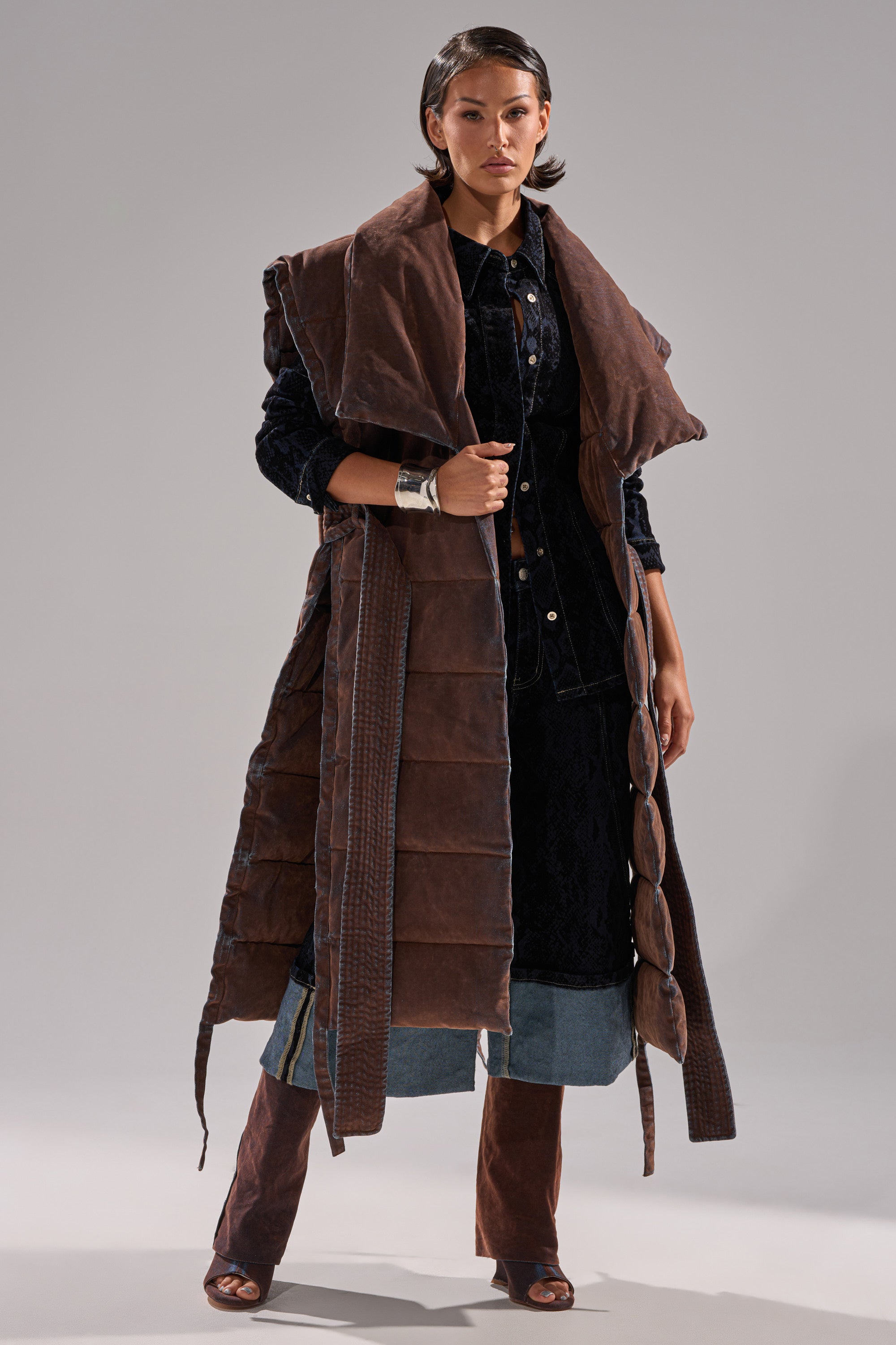 A woman stands confidently in the PILOT JONES LONGLINE PUFFER VEST IN VELVET DENIM over a dark denim dress and wide-leg jeans, accessorized with a chunky silver bracelet and open-toe brown shoes.