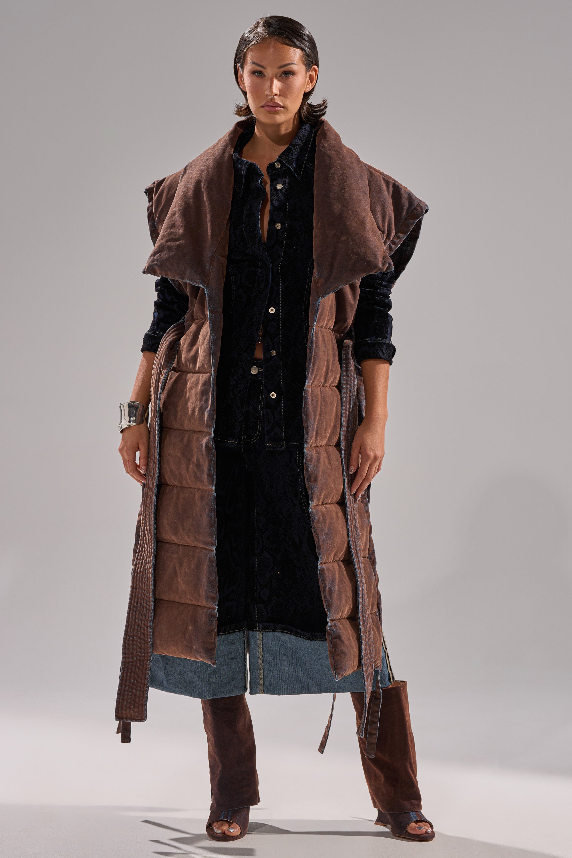 A woman stands against a plain background wearing the PILOT JONES LONGLINE PUFFER VEST IN VELVET DENIM over a dark button-up dress with denim details, paired with brown knee-high open-toe boots and a silver cuff bracelet.