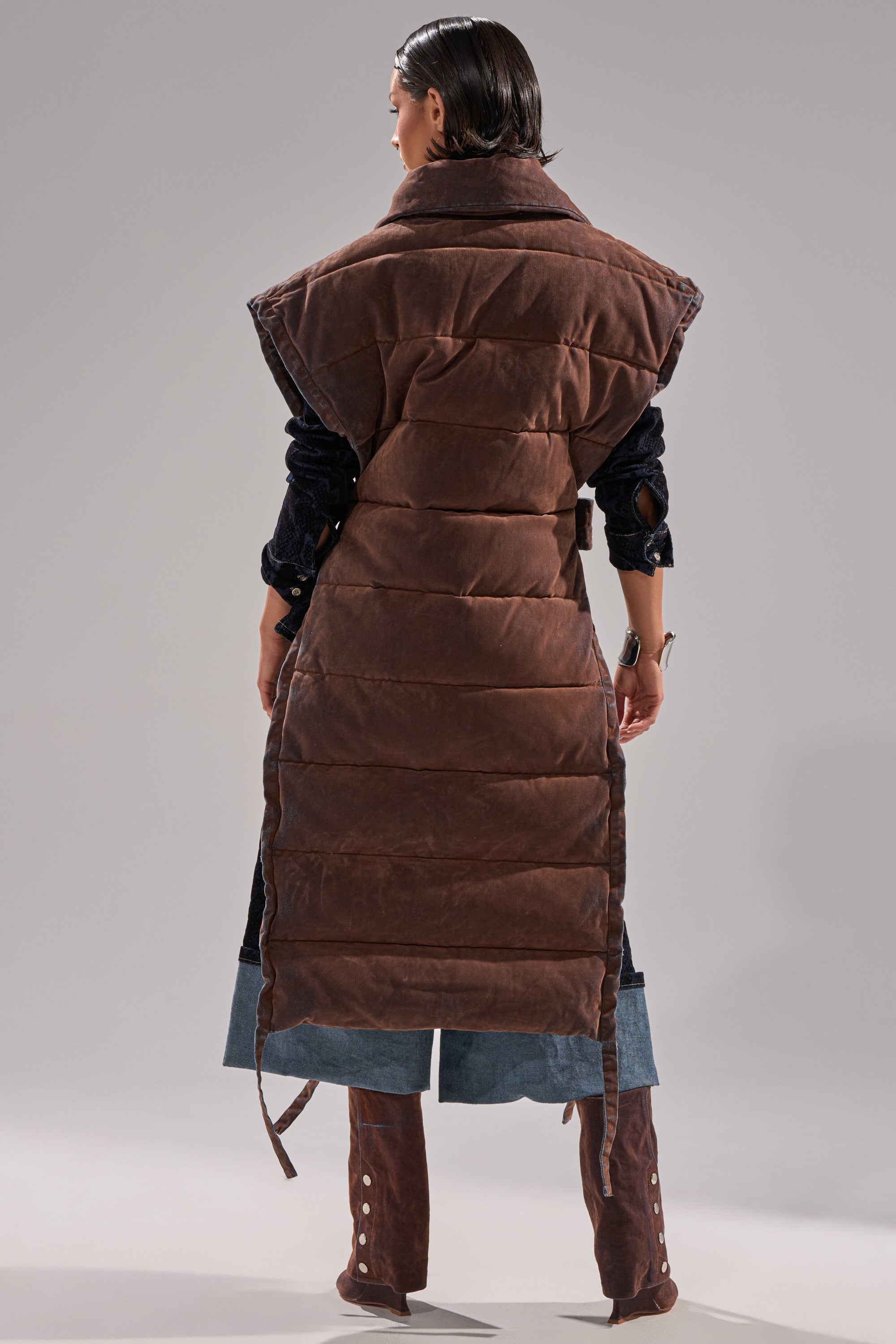 A person stands with their back to the camera, wearing the PILOT JONES LONGLINE PUFFER VEST IN VELVET DENIM over blue jeans; brown knee-high boots complete the look against a plain, light-colored background.