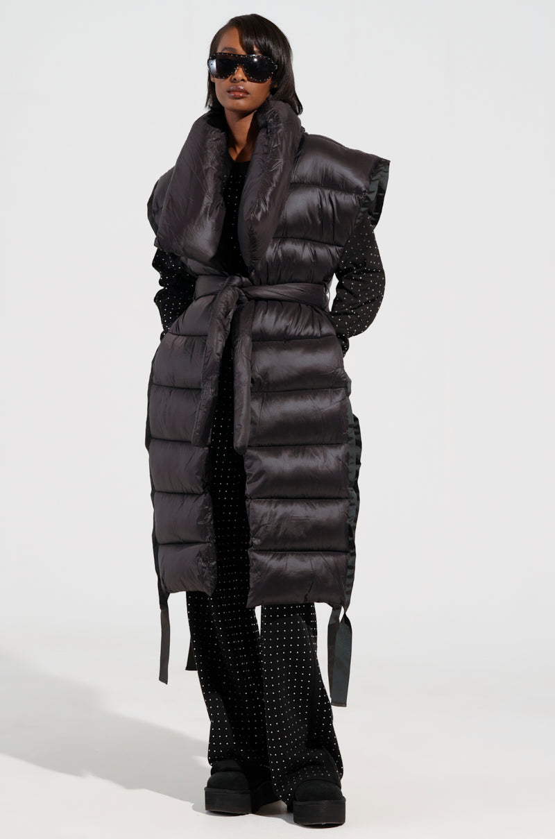 A person wearing sunglasses and the PILOT JONES LONG LINE PUFFER VEST, featuring a tie waist, over a black studded outfit. They stand with confidence against a plain background.