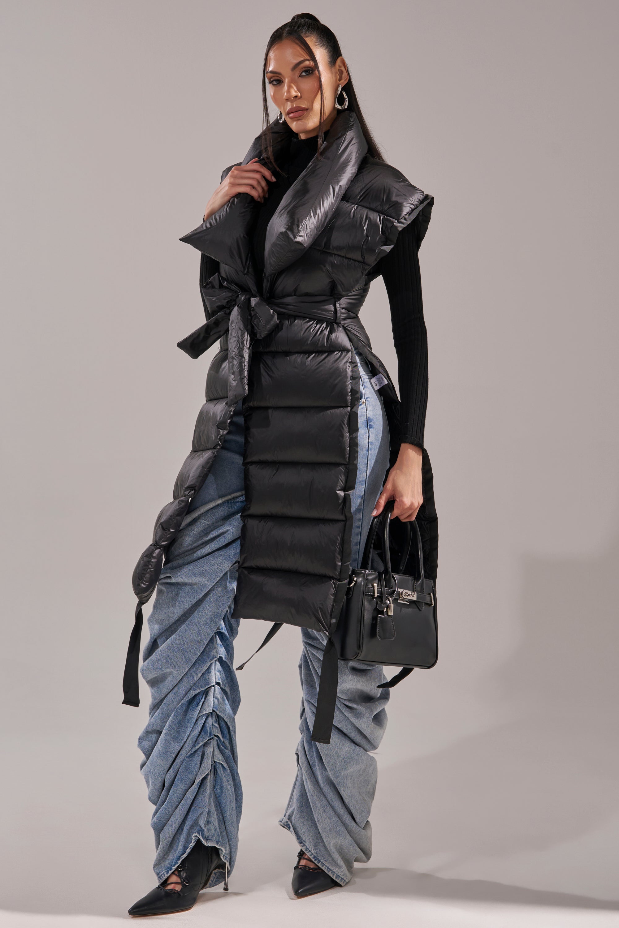 A woman poses in the PILOT JONES LONG LINE PUFFER VEST IN BLACK over a black turtleneck, styled with ruched wide-leg blue jeans, pointed black heels, hoop earrings, and a small black handbag against a plain background.
