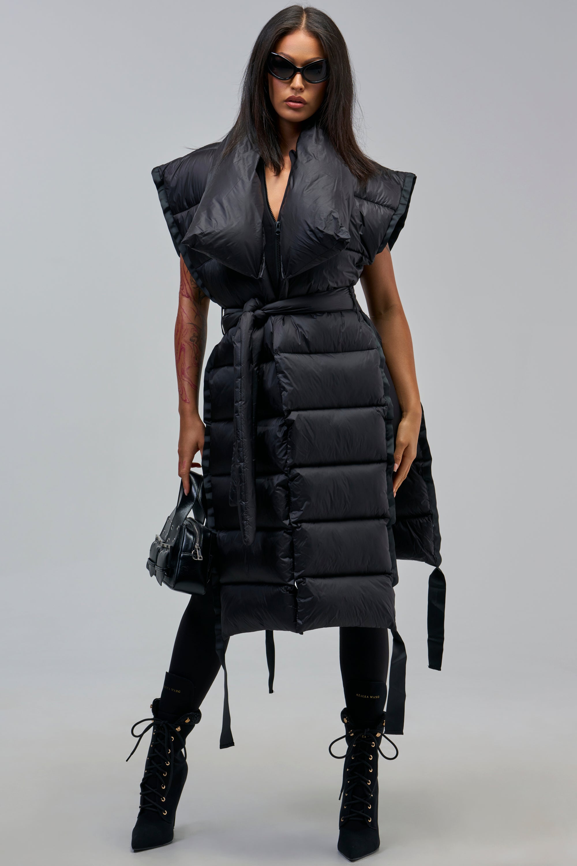 Confident woman wearing the PILOT JONES LONG LINE PUFFER VEST IN BLACK over leggings with lace-up boots, sunglasses, and a handbag, perfecting cold-weather layering against a gray backdrop.