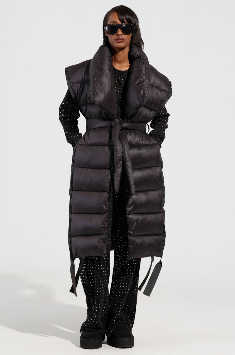 A person confidently stands wearing the PILOT JONES LONG LINE PUFFER VEST, featuring a tie waist and wide collar. They have on a black outfit with white polka dots, complemented by black sunglasses and sleek straight hair elegantly draped over their shoulders, creating an elegant longline silhouette.