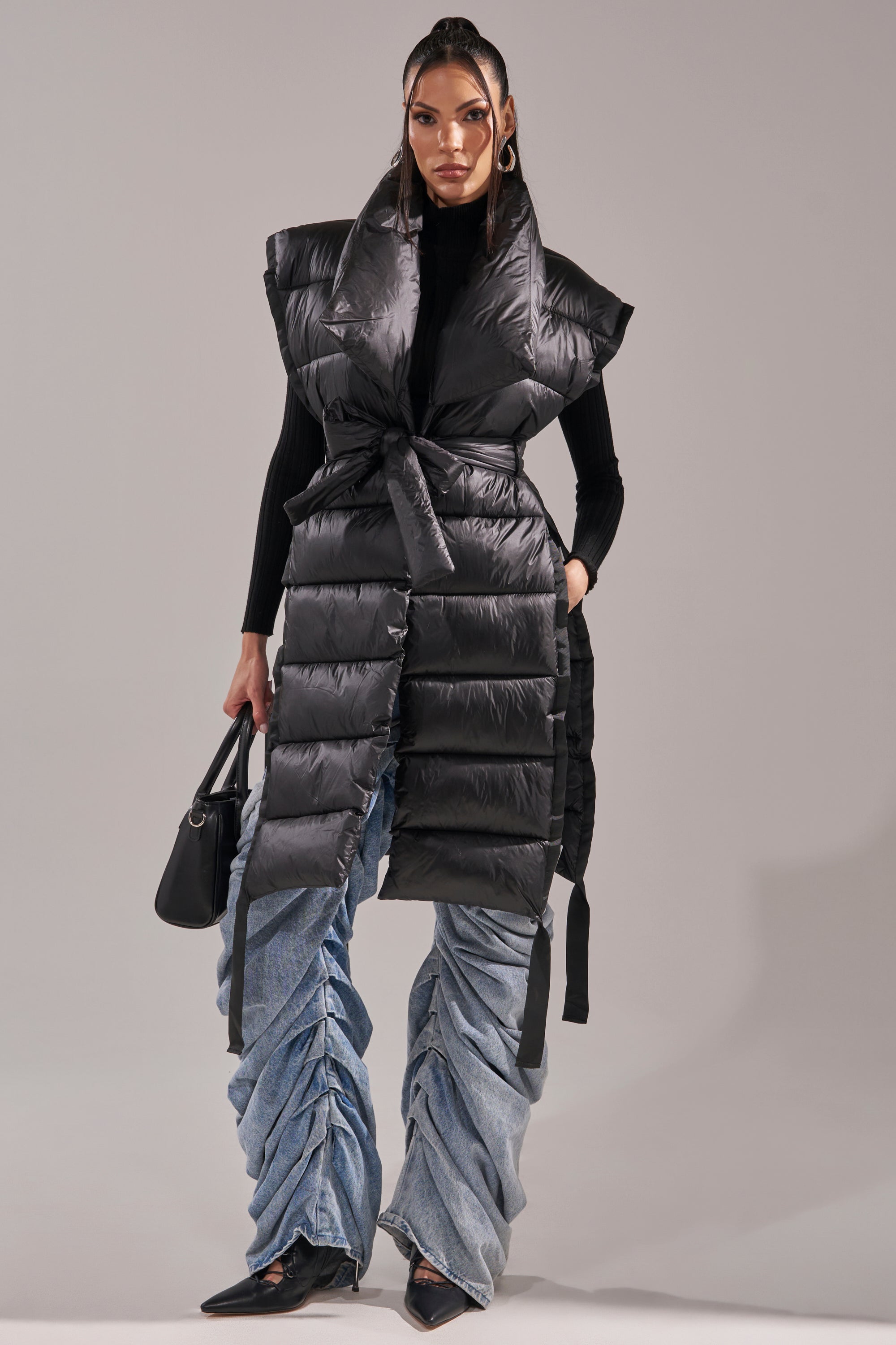 A woman models the PILOT JONES LONG LINE PUFFER VEST IN BLACK with a hood, layered over a black turtleneck, blue ruched jeans, and pointed shoes, carrying a black handbag against a plain gray background.