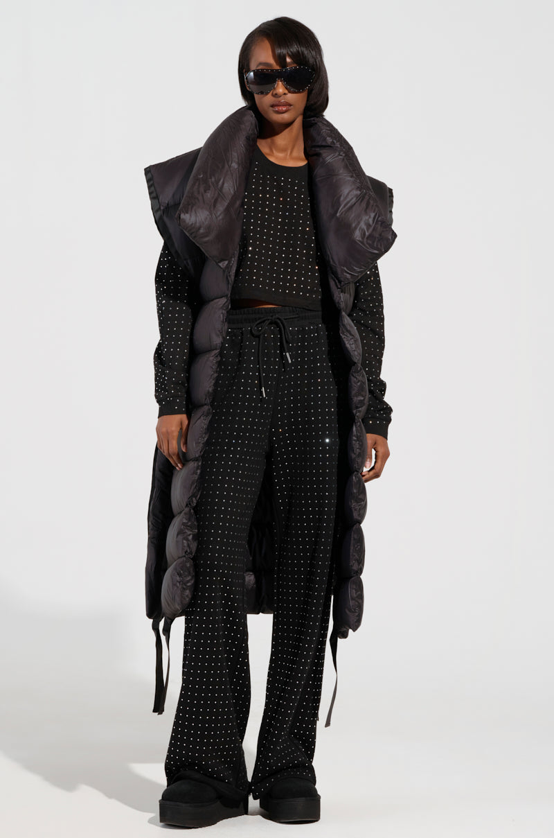 A person confidently poses in a chic black outfit adorned with silver dots, showcasing a cropped top, tie-waist wide-legged pants, and the PILOT JONES LONG LINE PUFFER VEST. They have accessorized with sunglasses and are standing against a plain white background.