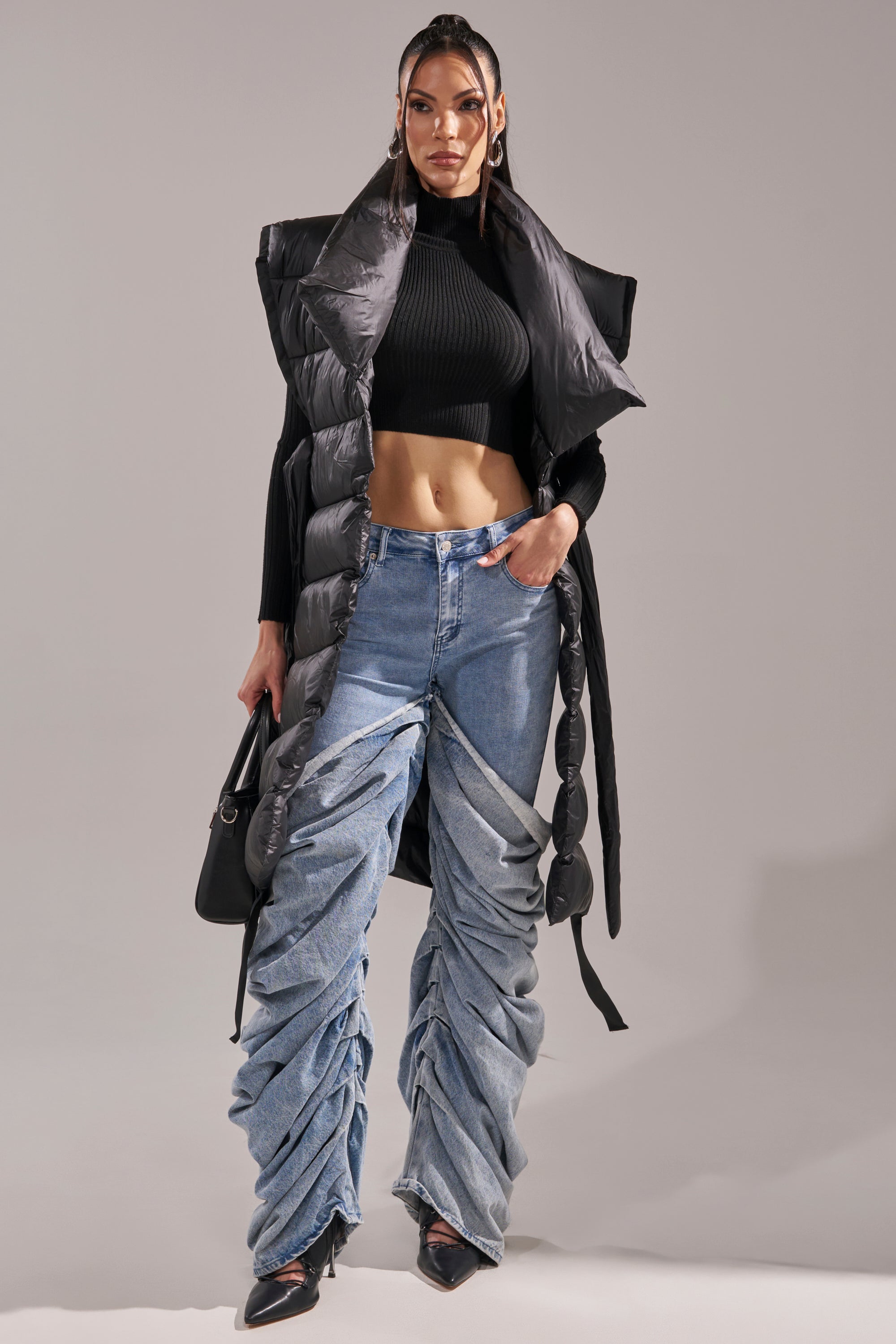 A woman stands confidently in the PILOT JONES LONG LINE PUFFER VEST IN BLACK over a black crop top, with ruched wide-leg jeans, pointed black heels, a black handbag, and sleek hair styled in two ponytails.