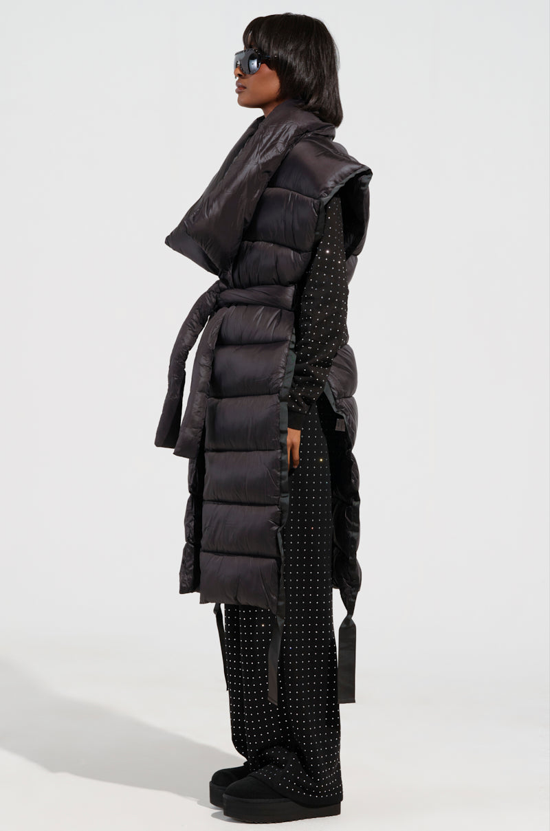 A person standing in profile showcases a striking silhouette with the PILOT JONES LONG LINE PUFFER VEST in black, featuring a tie waist and large collar. It is layered over a dotted black shirt and pants. They accessorize with chic black sunglasses and platform shoes, posing against a plain background.