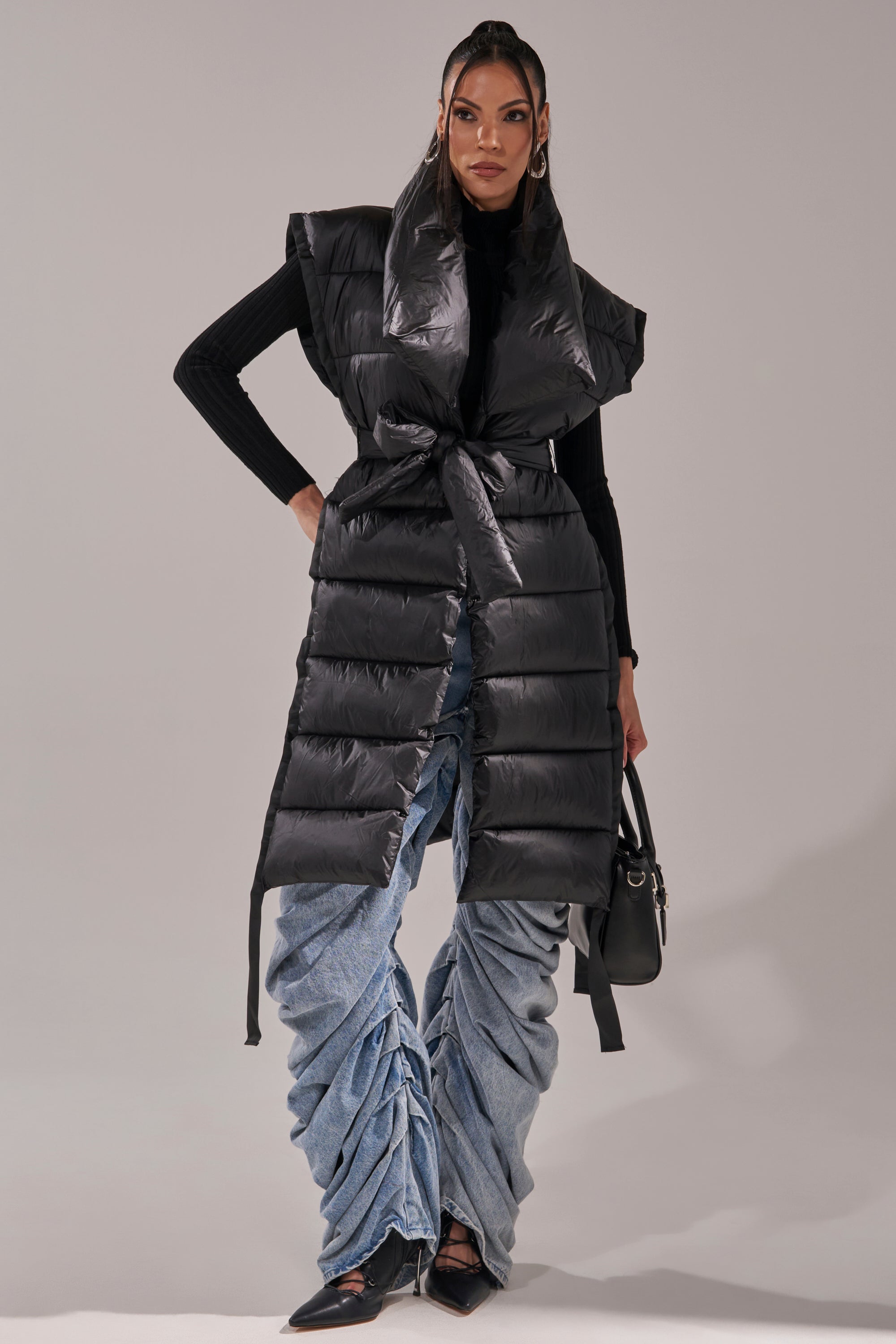 A woman models the PILOT JONES LONG LINE PUFFER VEST IN BLACK with a wide collar and waist tie, styled with a black turtleneck, ruched light blue jeans, black pointed shoes, and a small black handbag.