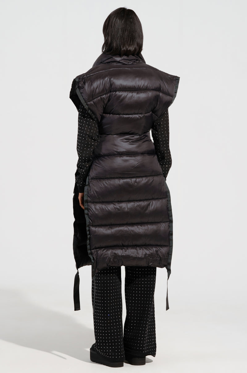 A person stands with their back to the camera, wearing the PILOT JONES LONG LINE PUFFER VEST in black over an outfit adorned with white polka dots. The vest has a sleeveless design, and the individual has dark hair, set against a plain white background.