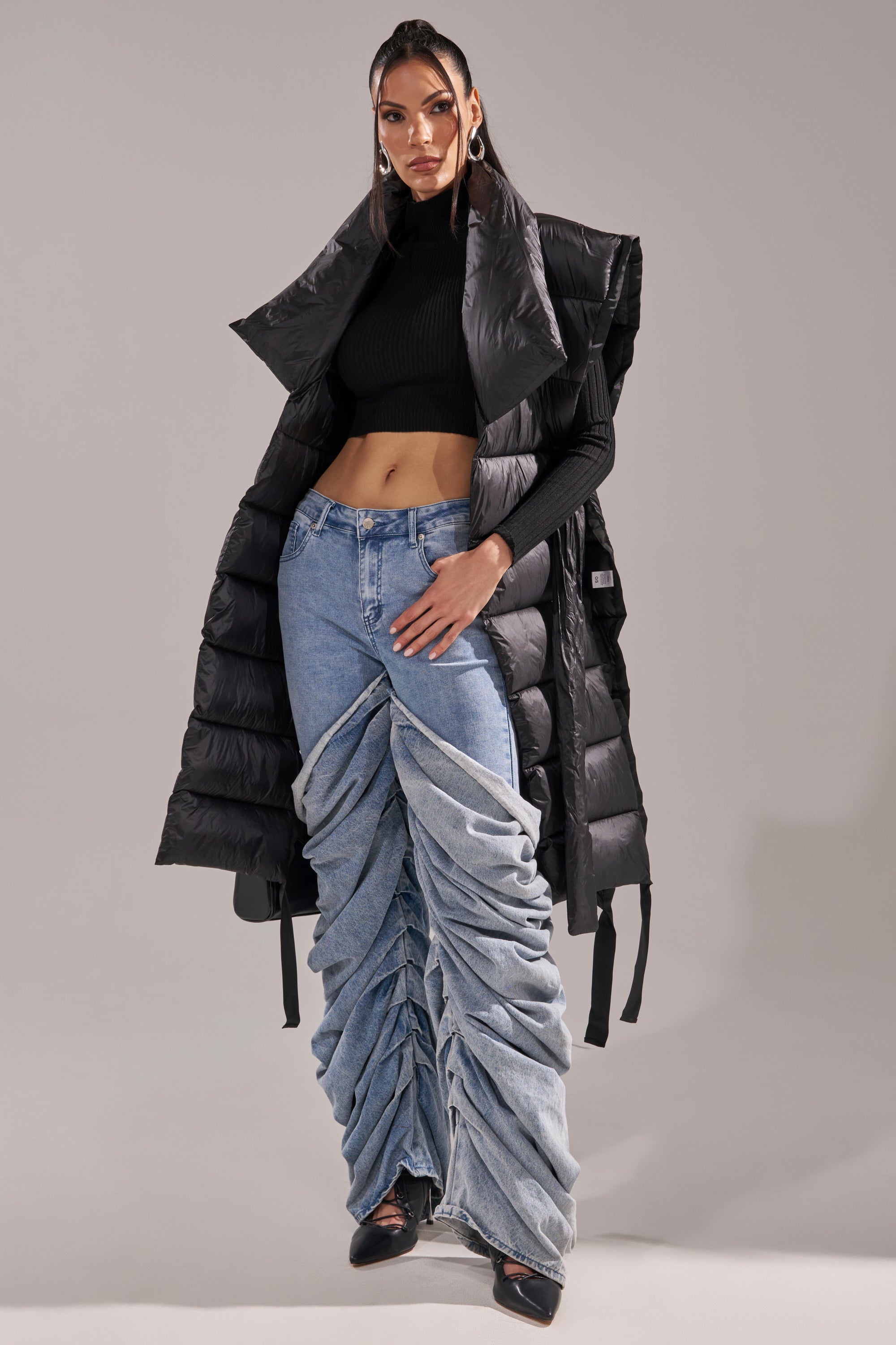 A woman poses in the PILOT JONES LONG LINE PUFFER VEST IN BLACK over a cropped turtleneck with ruched, oversized denim jeans. She completes the look with black heels and hoop earrings, standing against a light gray background.