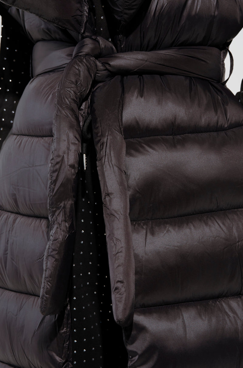 A close-up of the PILOT JONES LONG LINE PUFFER VEST in black reveals its shiny, quilted texture and tie waist. The vest's longline design exudes elegance, with glimpses of dotted fabric visible beneath it.