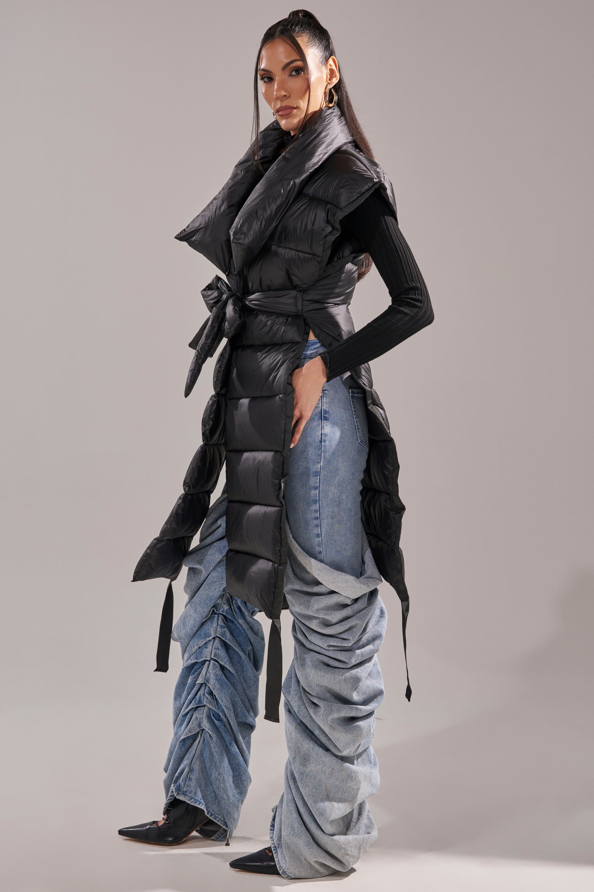A woman poses in the PILOT JONES LONG LINE PUFFER VEST IN BLACK with a belt over a black turtleneck and baggy ruched blue jeans, paired with black pointed shoes, against a plain gray background.