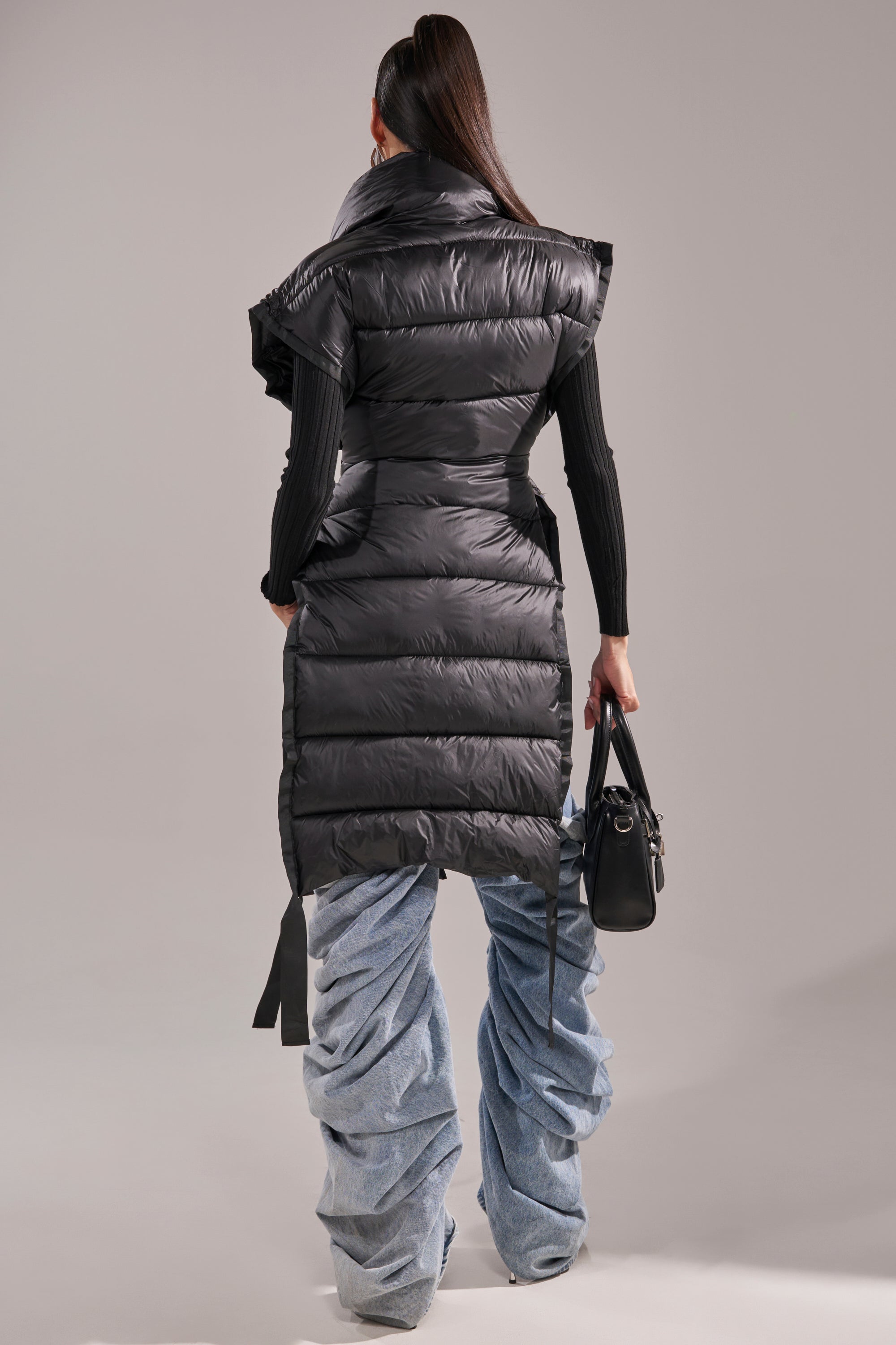 A person with long dark hair in a ponytail faces away, wearing the PILOT JONES LONG LINE PUFFER VEST IN BLACK over a black top and ruched light blue jeans, holding a black handbag.