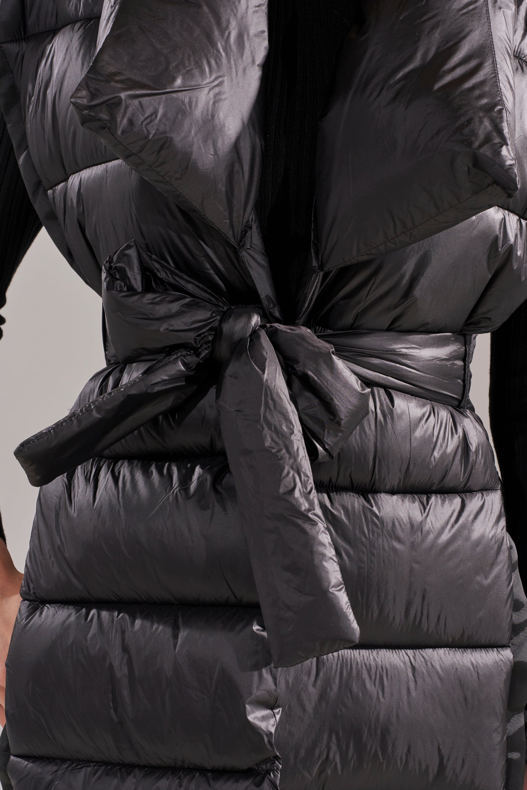Close-up of the PILOT JONES LONG LINE PUFFER VEST IN BLACK, featuring a shiny finish, horizontal quilting, and a tie belt at the waist, with part of a black ribbed sleeve visible.